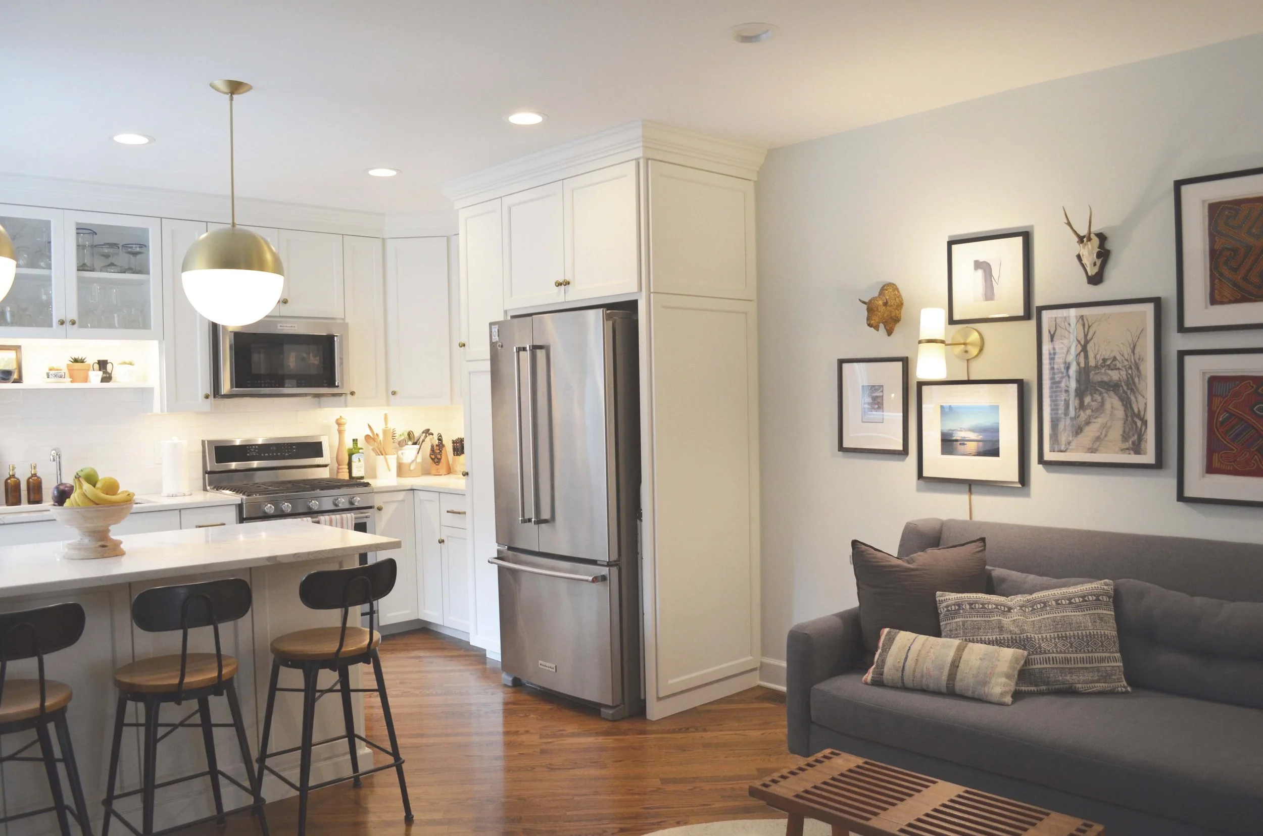 Open-concept kitchen with white cabinets, stainless steel appliances, black bar stools, and a gray sofa with decorative pillows, all in a cozy living space.