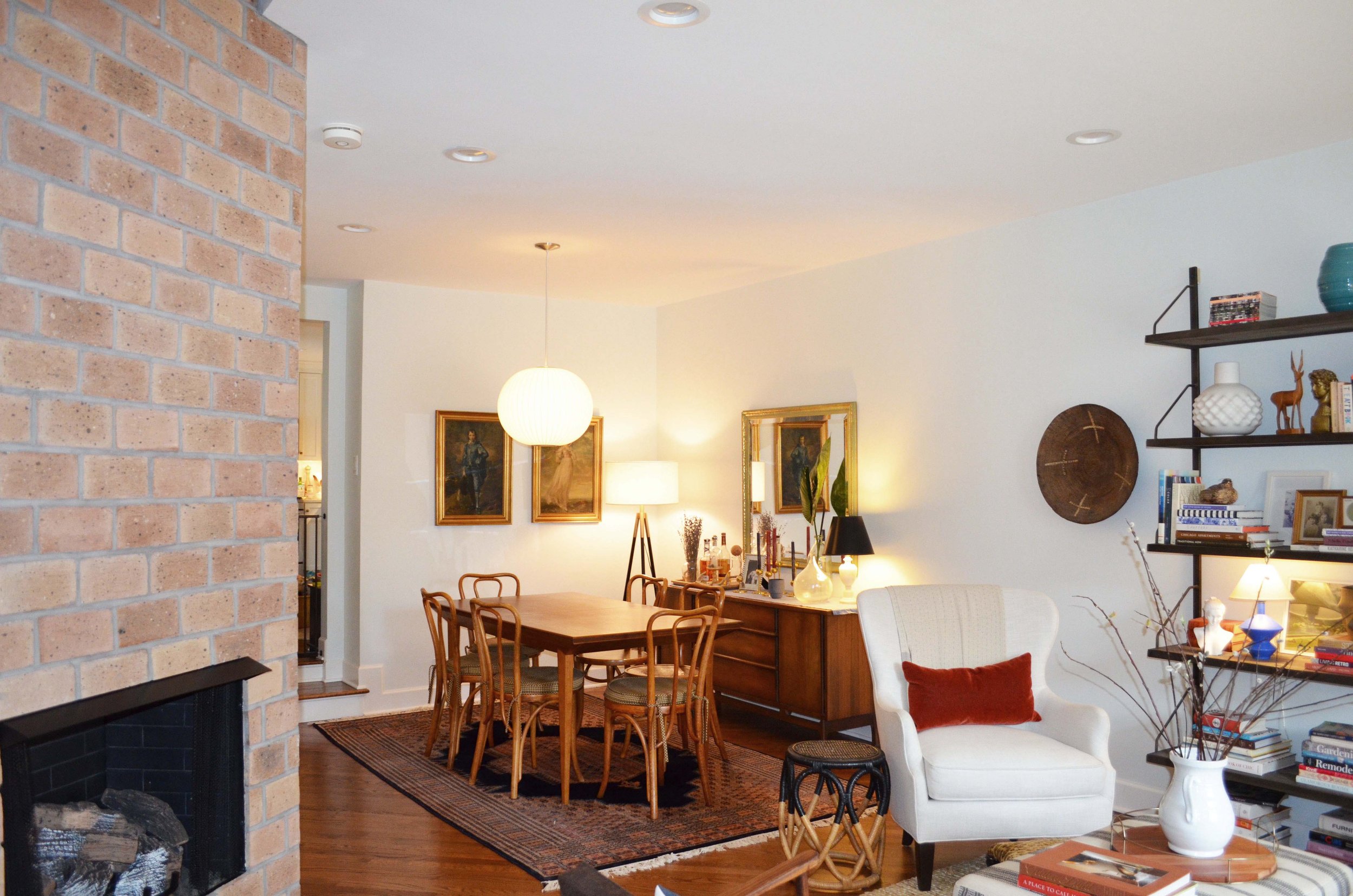 A dining room with a wooden table and six chairs, a sideboard with lamps and decorative objects, framed paintings on the wall, a white armchair with a red pillow, a shelf with books and decorative items, a brick fireplace, and various lamps providing