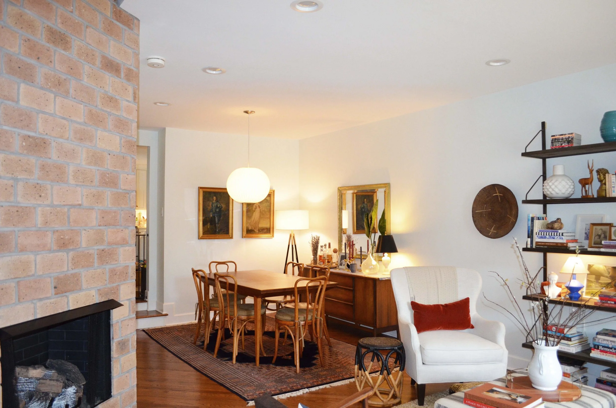 A cozy dining and living area with a brick fireplace on the left, a wooden dining table with six chairs, and a white armchair with a red pillow. Decor includes framed paintings, a large mirror, various lamps, and shelves with books and decorative obj
