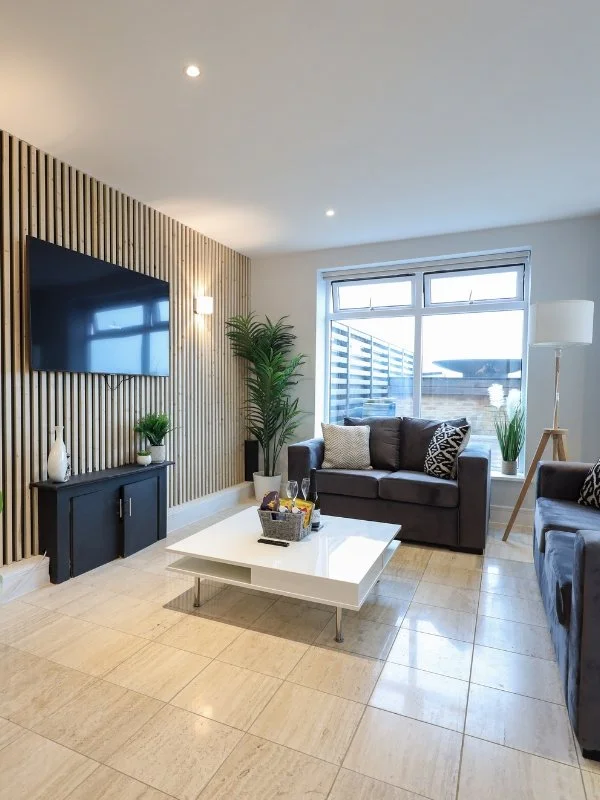 Living room with a flat-screen TV on a wooden accent wall, black entertainment unit, potted plant, gray sofas with patterned cushions, white coffee table with snacks and drinks, large window with blinds, and a floor lamp.