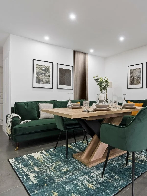 Modern dining room with green velvet sofa, matching green chairs, wooden dining table with tableware, decorative rug, and black and white framed art on white walls.