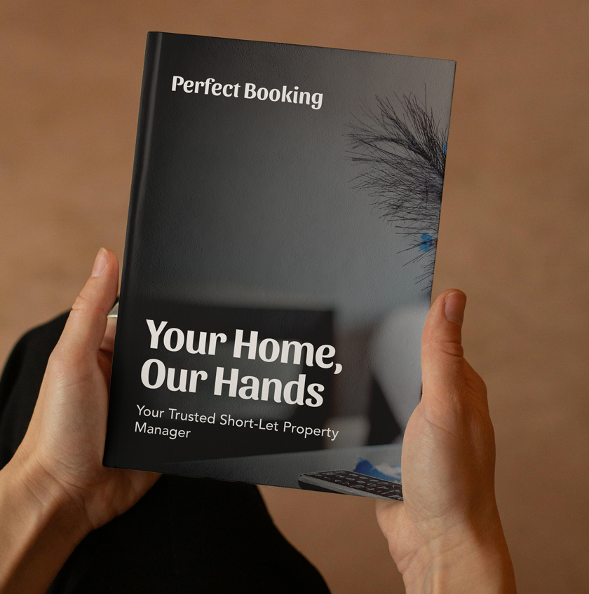 A person holding a black book titled 'Perfect Booking' with a subtitle 'Your Home, Our Hands' and a tagline 'Your Trusted Short-Let Property Manager.' The cover features a close-up of a white ceramic cup, a blue pen, and a TV remote, with a blurred brown background.