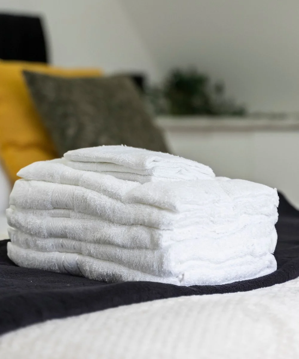 Stack of neatly folded white towels on a black and white bedspread in a bedroom with pillows and a plant in the background.