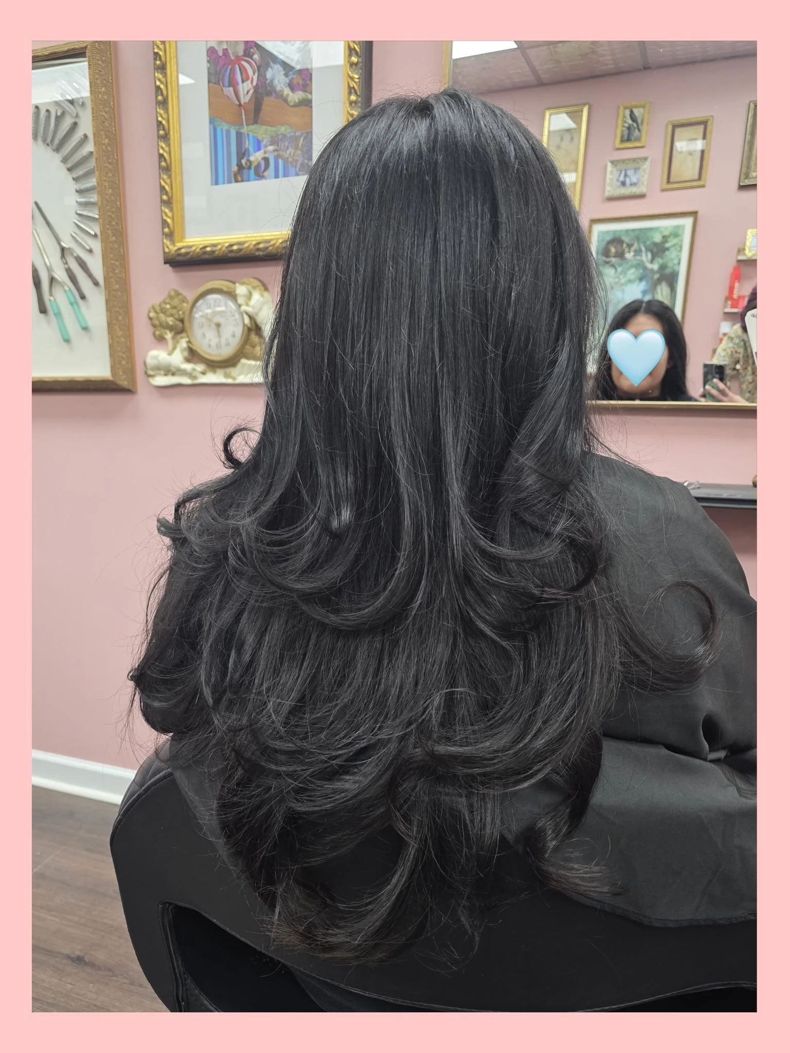 Back view of a woman with long, layered, black hair with soft curls at the ends, sitting in a salon chair in front of a mirror. The salon has pink walls decorated with framed artwork and a clock.