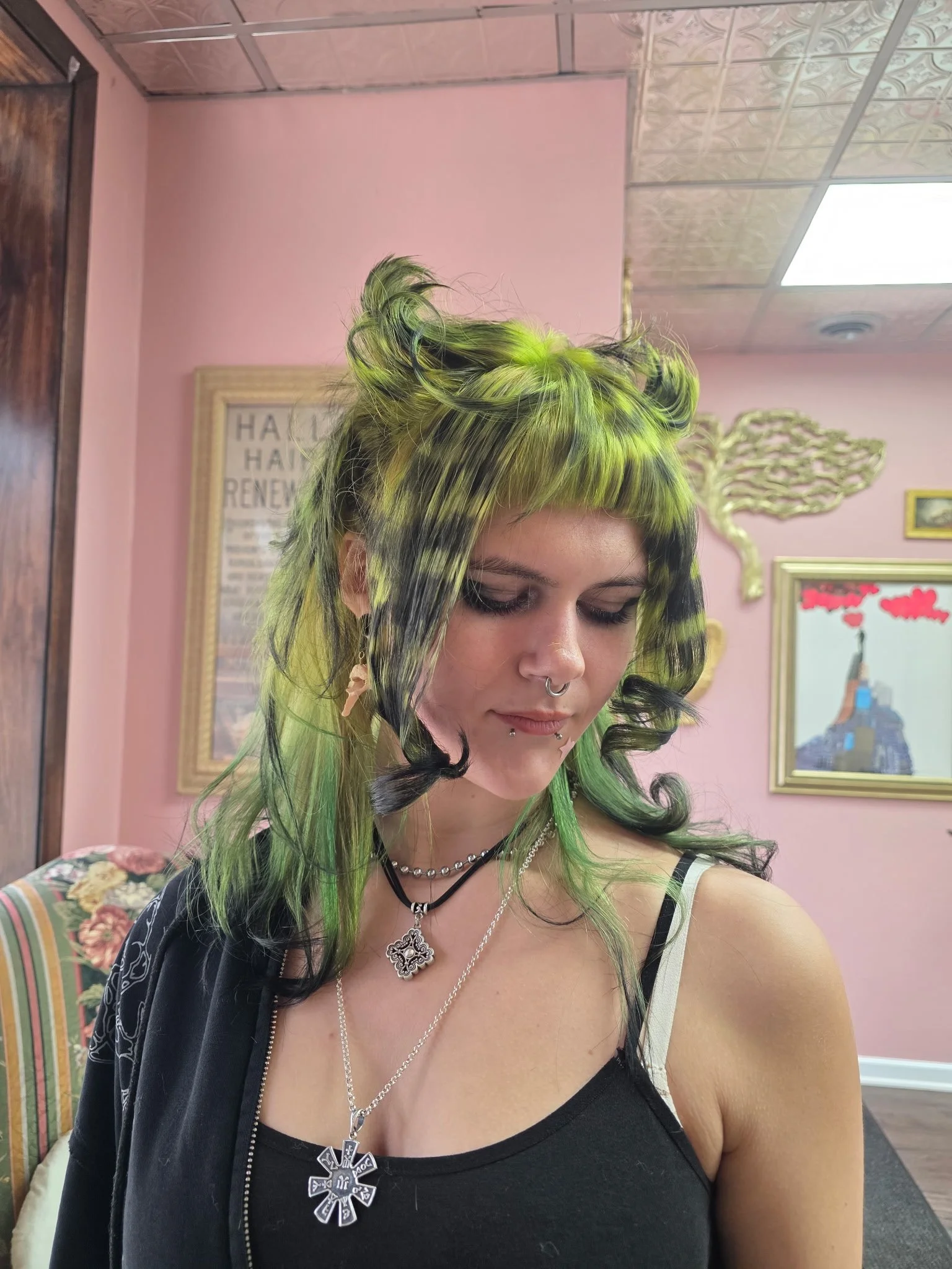 Young woman with green and black styled hair, multiple piercings, wearing layered necklaces, standing indoors with pink walls and framed artwork in the background.