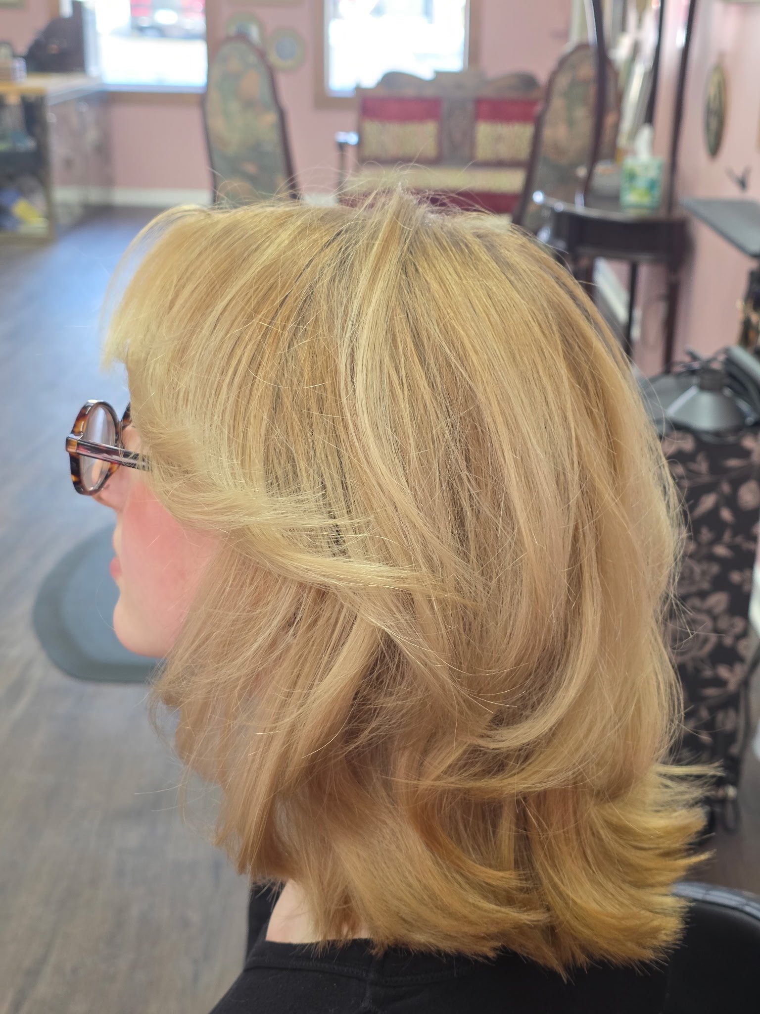 Side view of a woman with shoulder-length, layered blonde hair with soft waves, wearing glasses, in a hair salon.