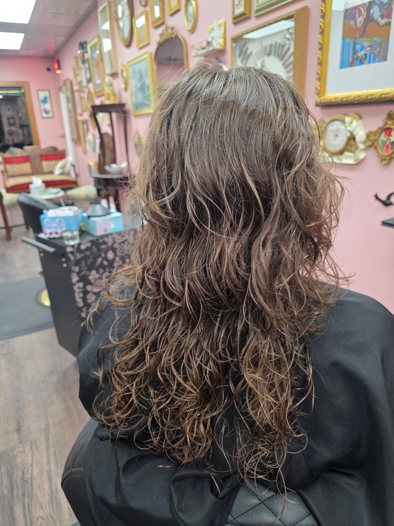Back view of a woman with long, wet, curly brown hair sitting in a room with pink walls decorated with various framed pictures and mirrors.