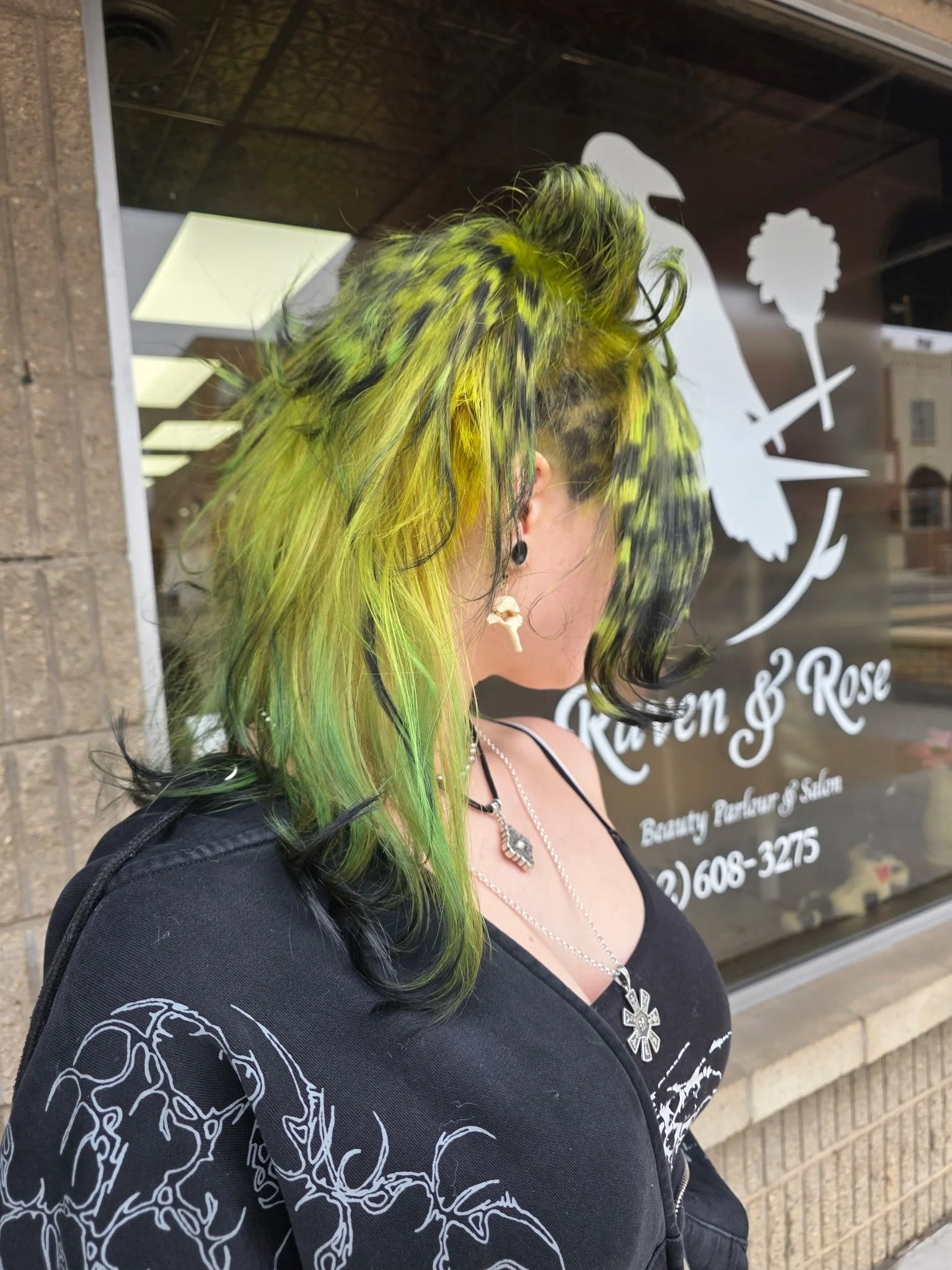 Person with vibrant green and black hair styled in a mohawk, wearing earrings and necklaces, standing outside a beauty salon with a floral logo on the window.
