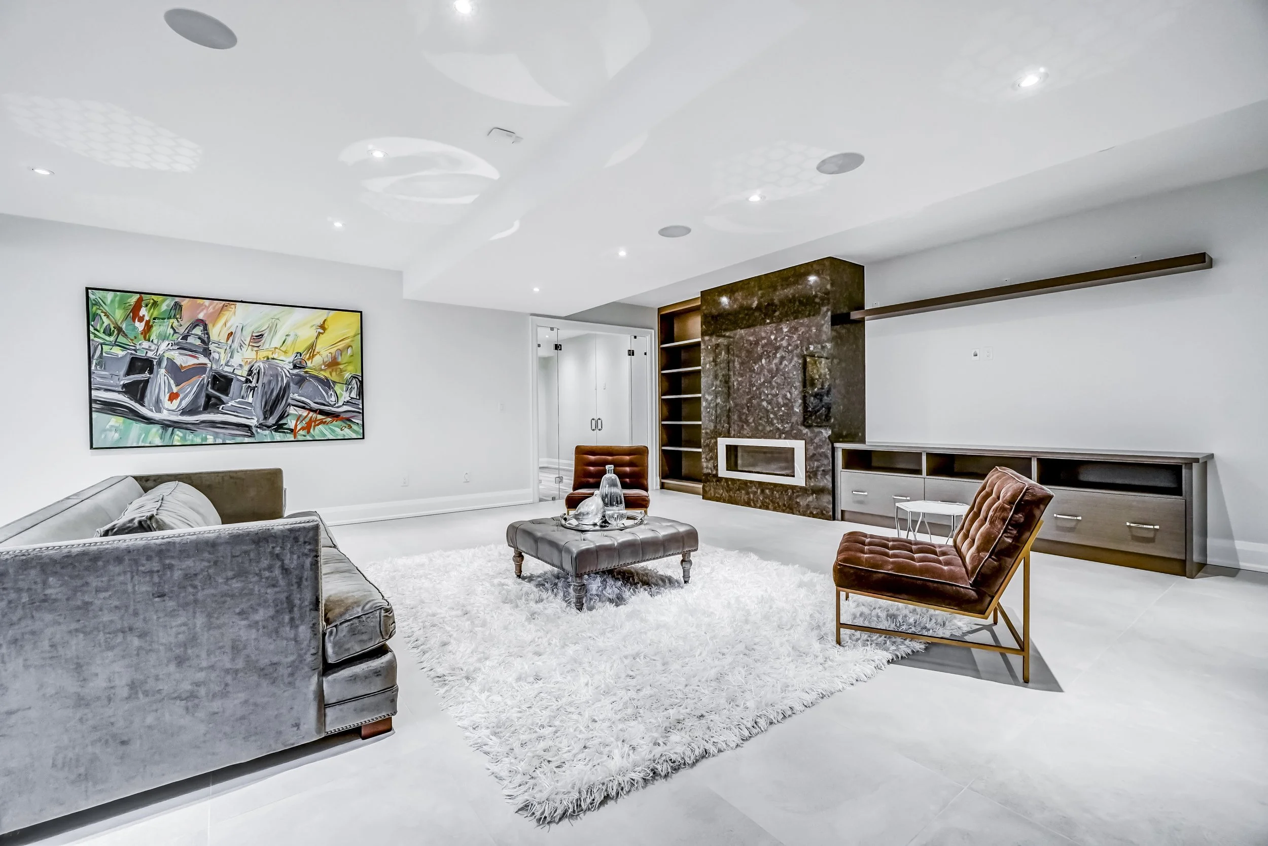 Modern living room with white walls, a fluffy white rug, a gray velvet sofa, brown leather chairs, a small side table, a large abstract painting, and a built-in fireplace with dark marble around it.
