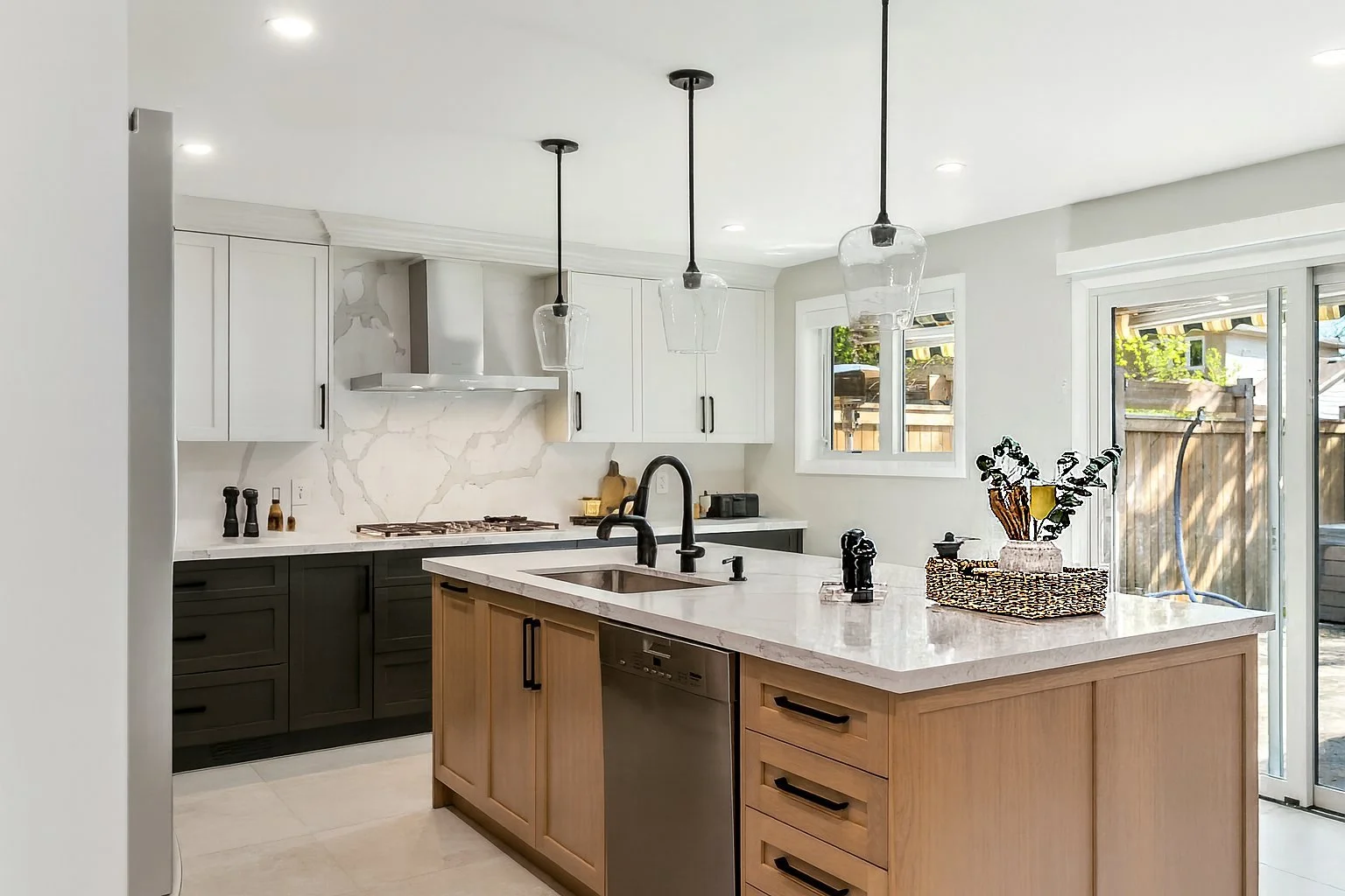 Modern kitchen with white upper cabinets, black lower cabinets, a marble backsplash, and a wooden kitchen island with a beige marble countertop. Pendant lights hang above the island, and there is a gas stove, a sink with a black faucet, and a dishwasher integrated into the island. Large windows and sliding glass door let in natural light.