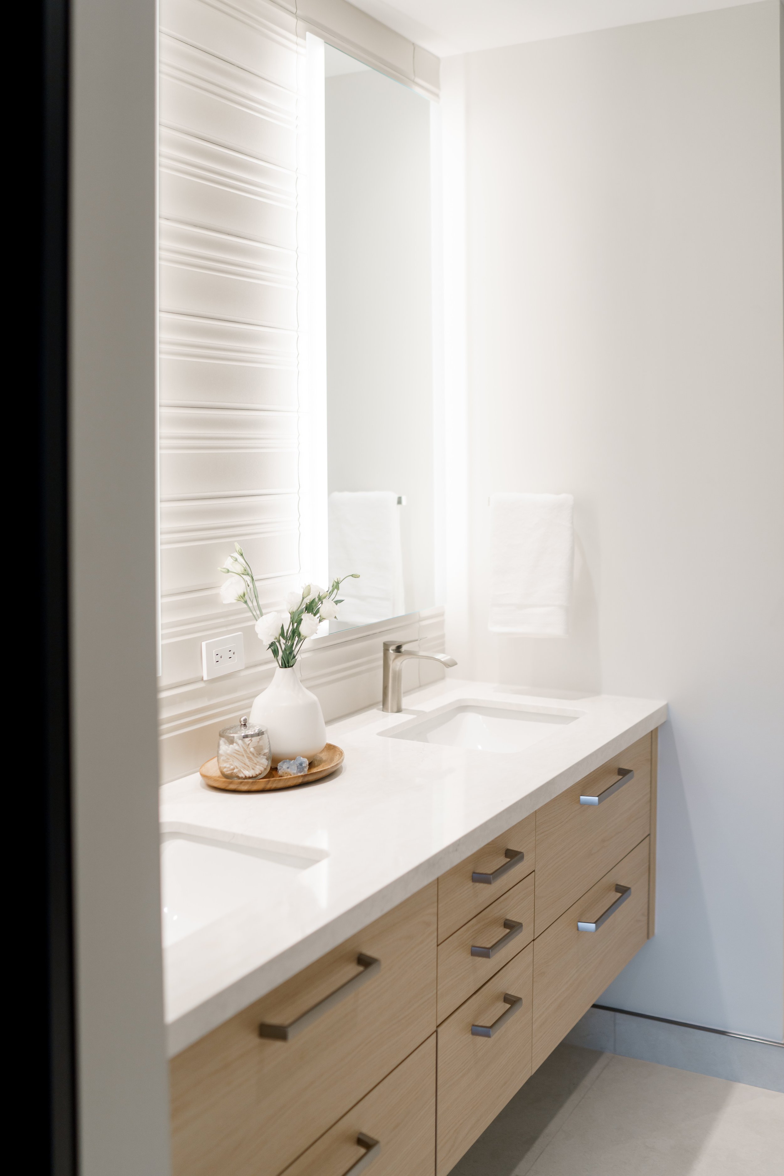 A modern bathroom vanity with a light wood finish and a white countertop. There is a single sink with a stainless steel faucet, a large mirror above the sink, and a vase with white flowers on a wooden tray. Towels are hanging on the wall, and there is a power outlet near the mirror.