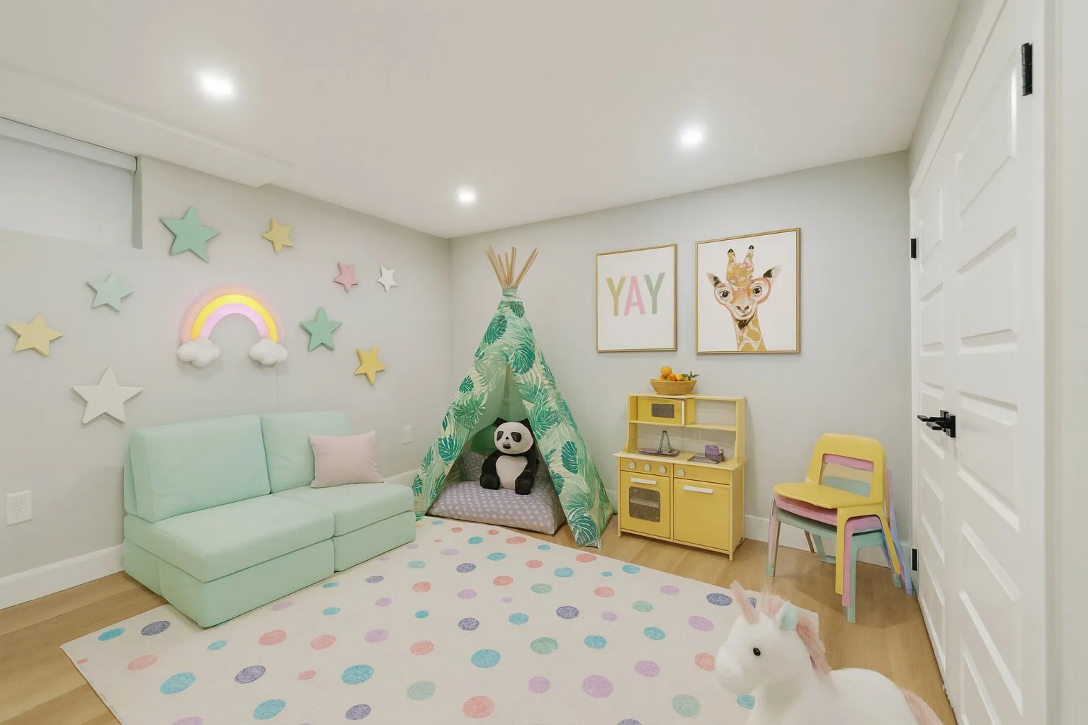 Children's playroom with pastel-colored decor, star and rainbow wall art, a mint green sofa, a small yellow toy kitchen, a unicorn plush toy, a teepee tent with a panda plush inside, and pastel chairs, with a white plush unicorn toy on the floor.