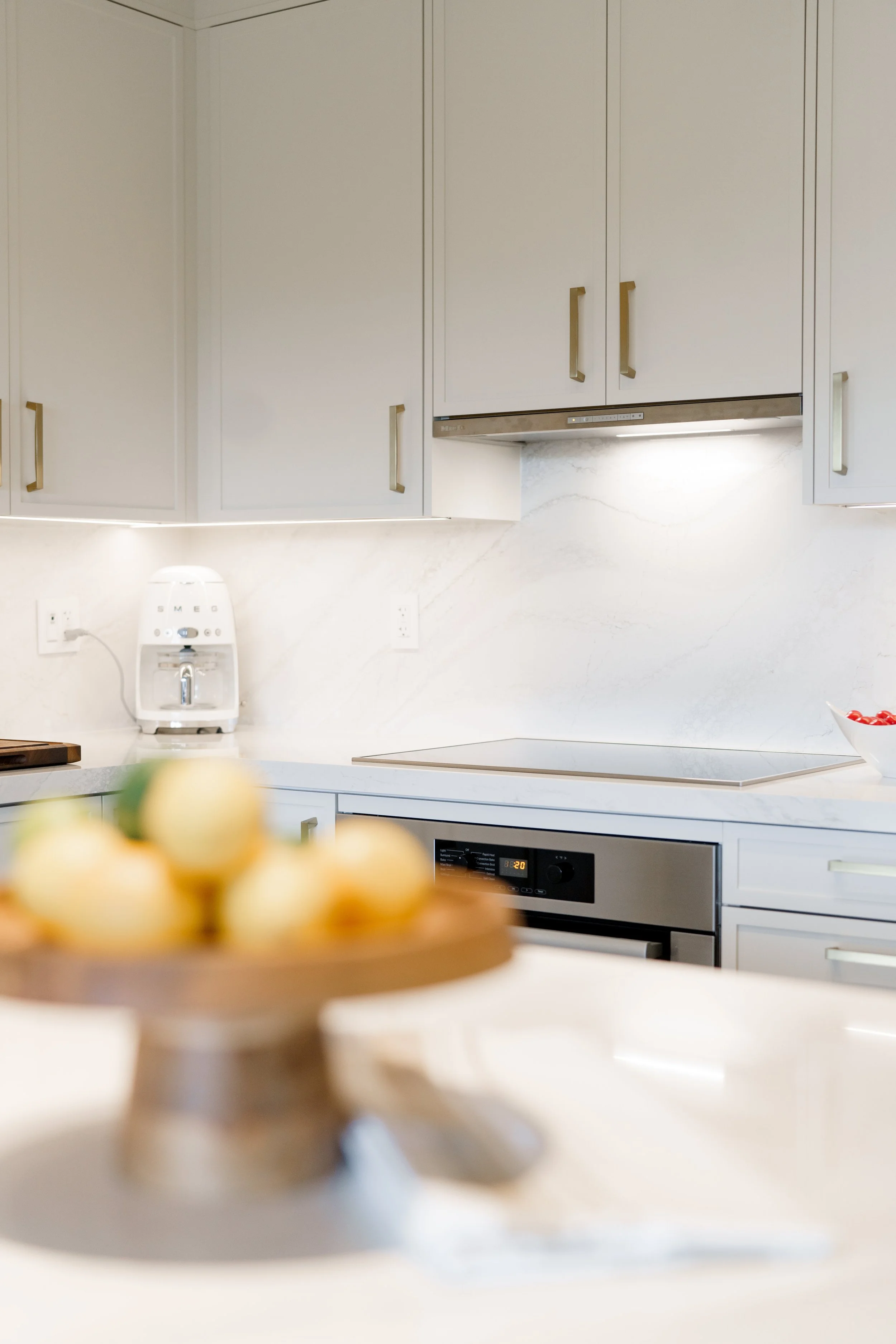 Modern kitchen with white cabinets, marble backsplash, and built-in stainless steel oven, with a bowl of cherries on the counter.