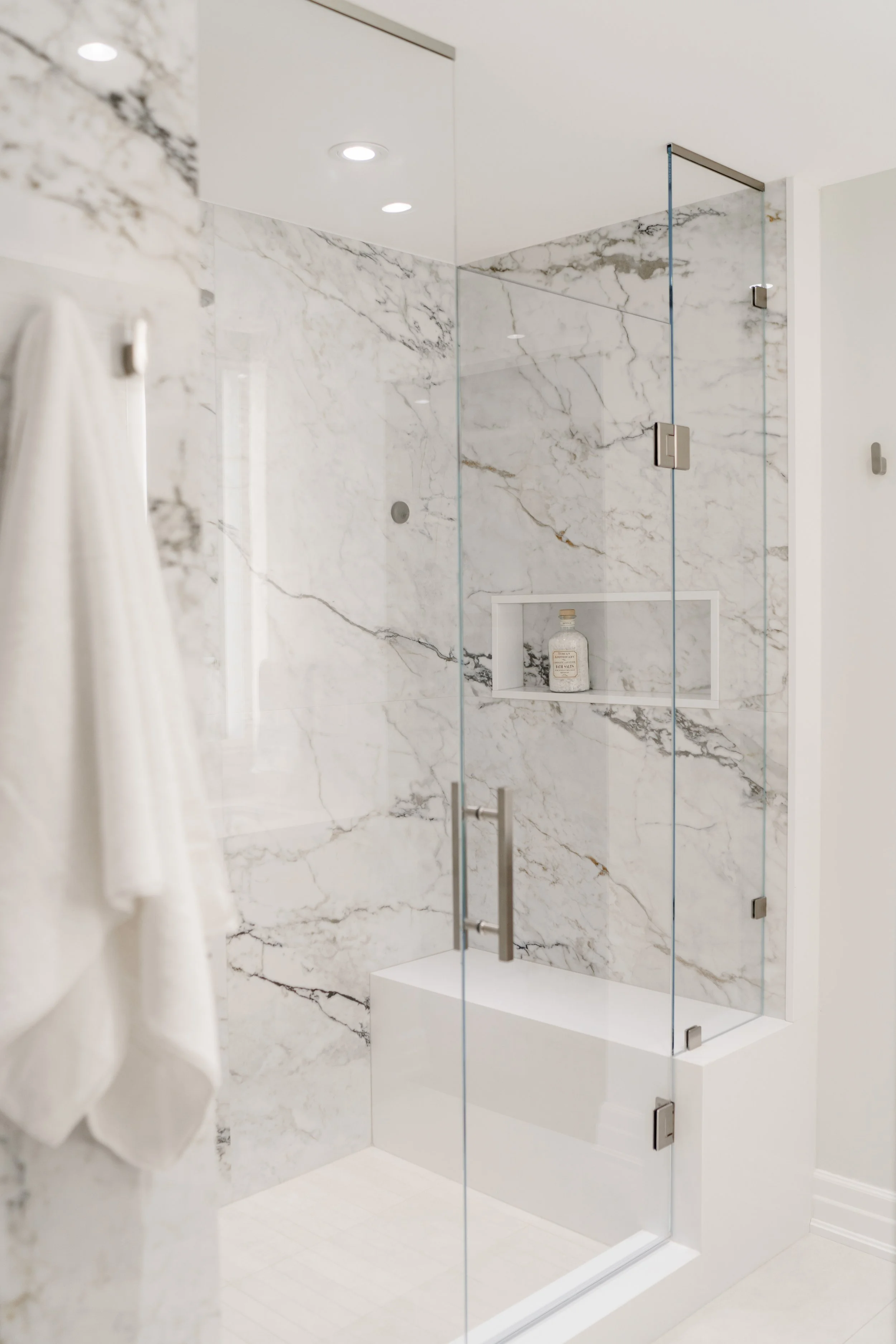 A modern glass-enclosed shower with white marble walls featuring gray veining, a built-in niche with a bottle of bathing product, and a sleek door handle.