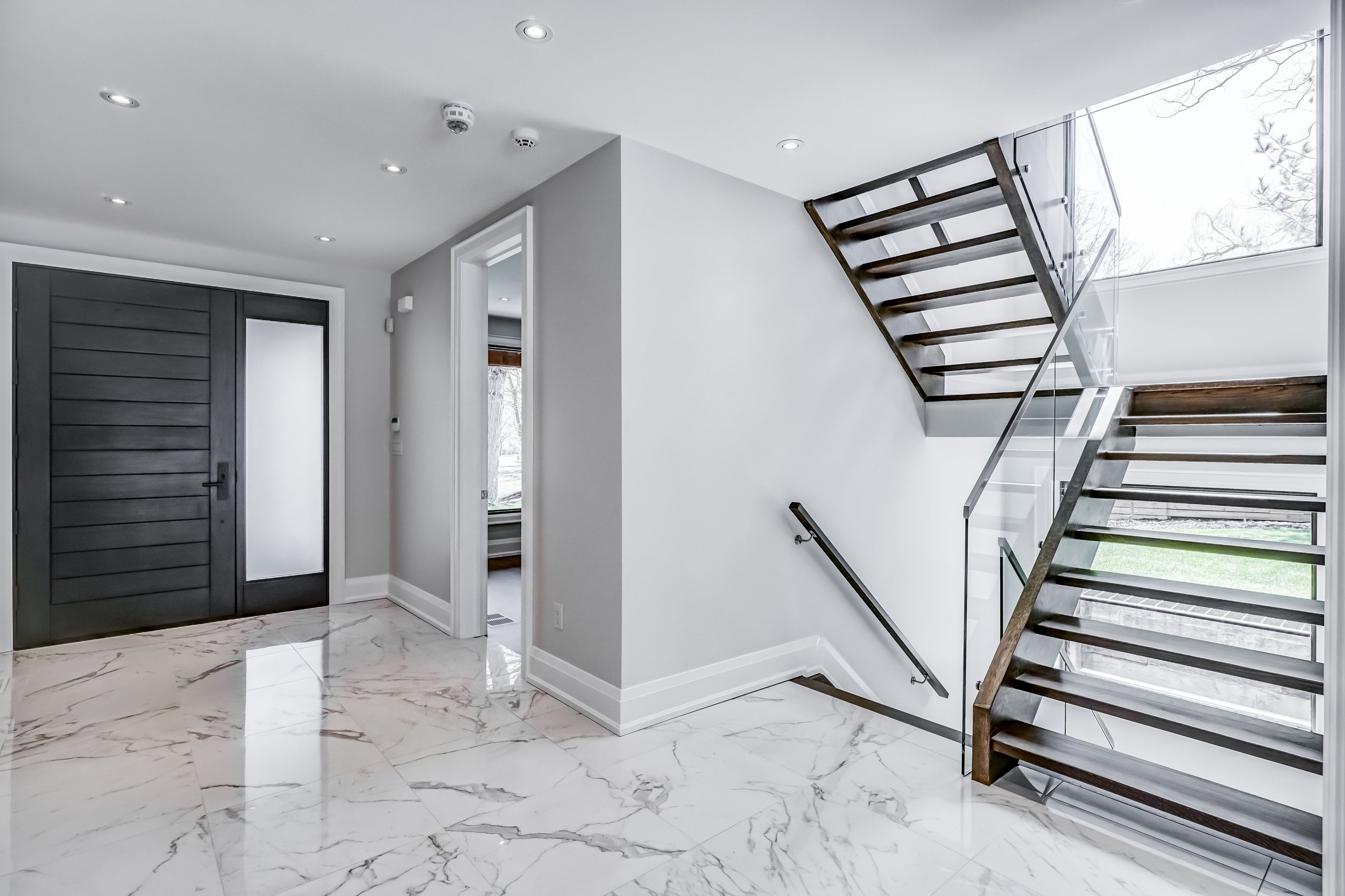 Modern staircase with wooden steps and glass railing in a bright open space with marble flooring and large window.