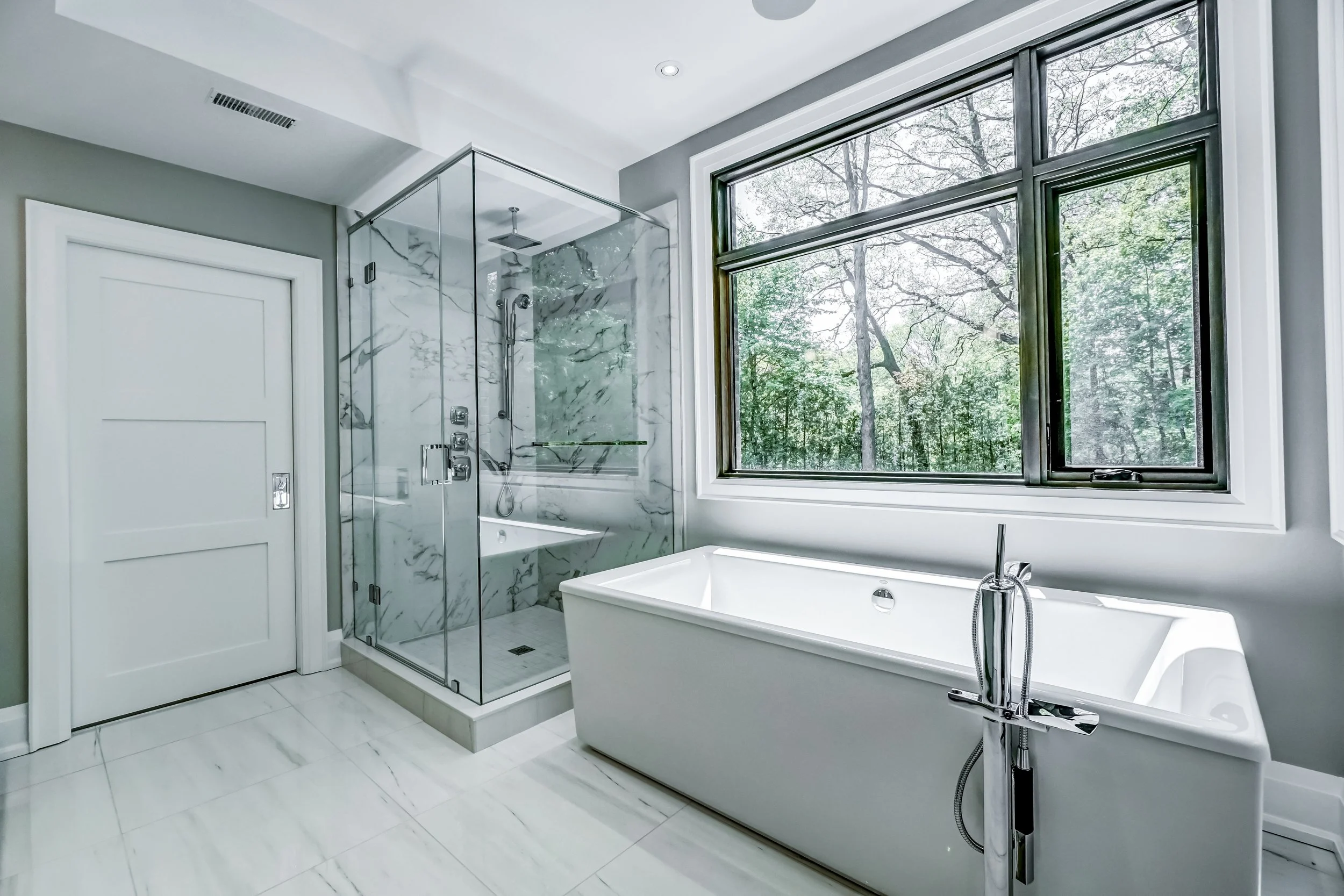 Modern bathroom with a large window overlooking trees, a glass-enclosed shower with marble walls, and a white freestanding bathtub.