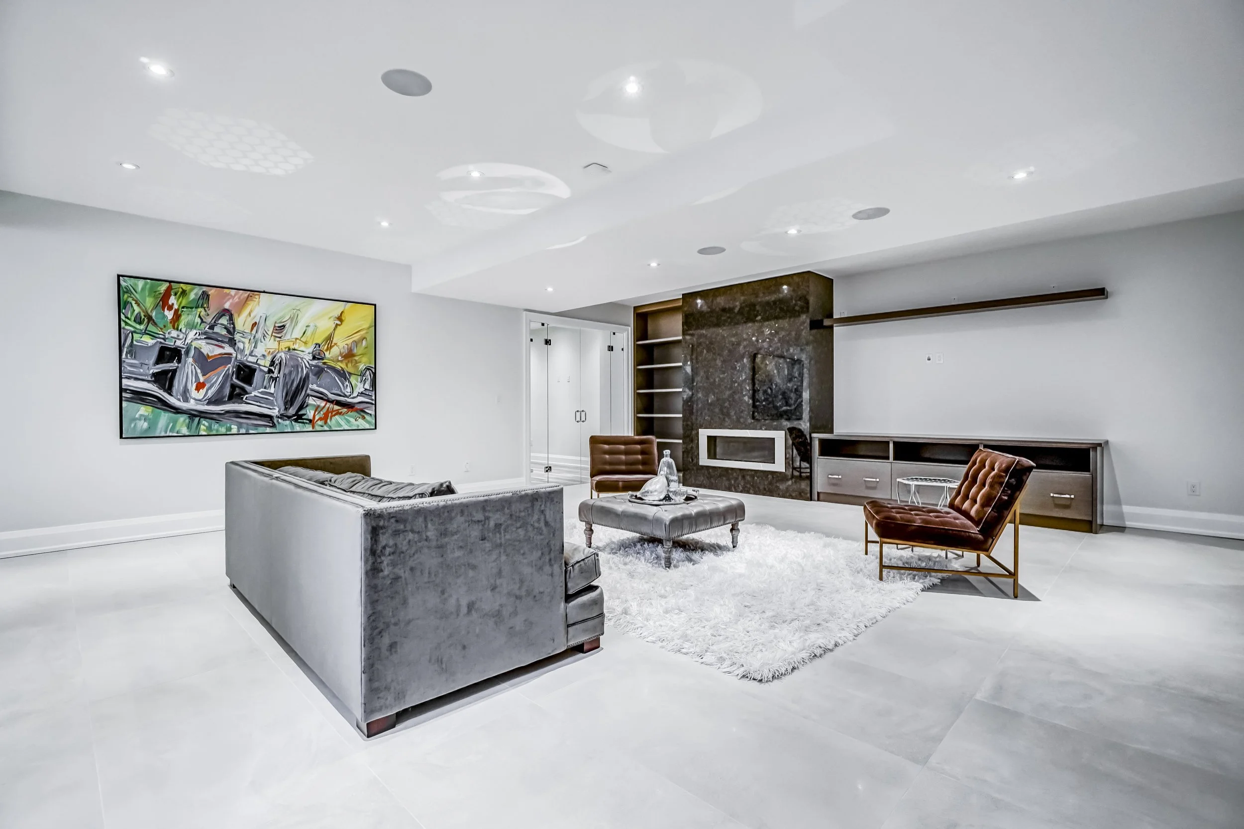 Modern living room with white walls, a gray velvet sofa, a brown leather chair, a stone fireplace, colorful abstract artwork on the wall, and a white shag rug.