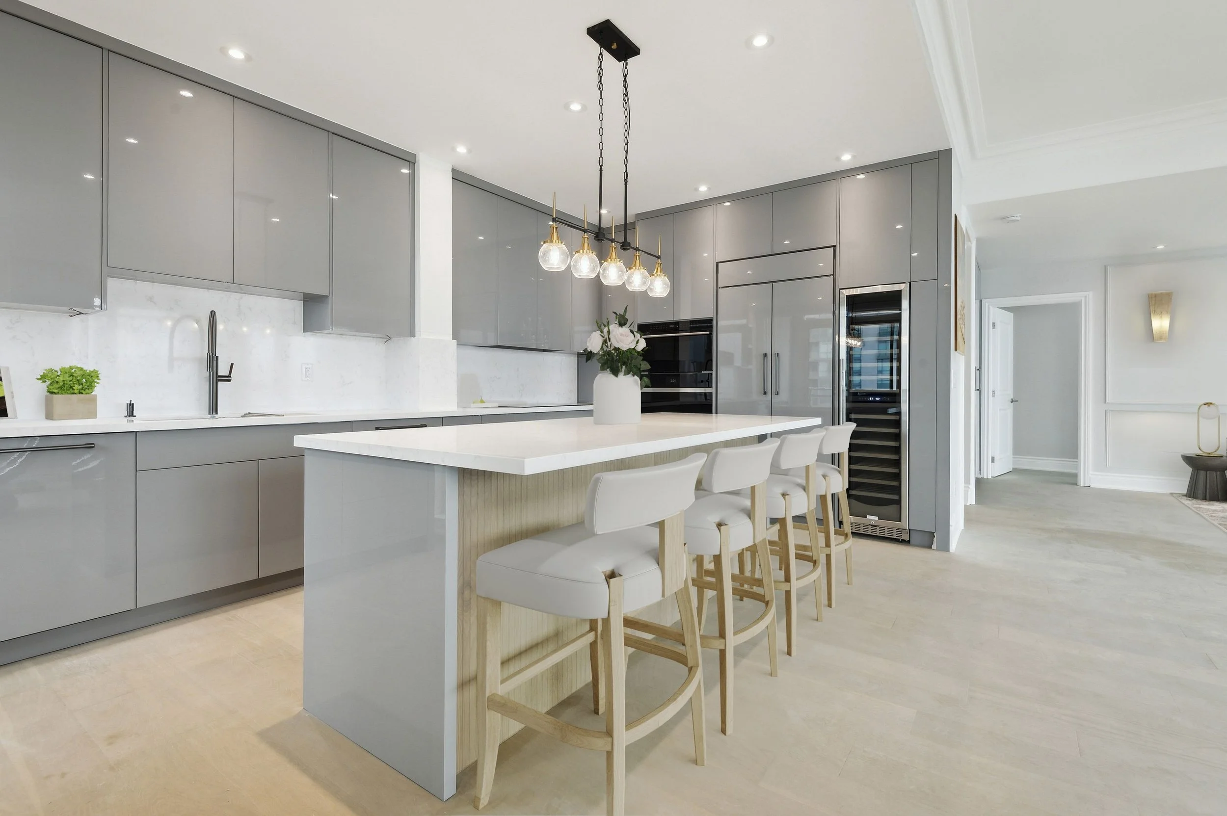 Modern kitchen with gray cabinets, white marble backsplash, island with white countertop, and four white barstools. Overhead lighting with five glass bulbs, and a vase of flowers on the island.