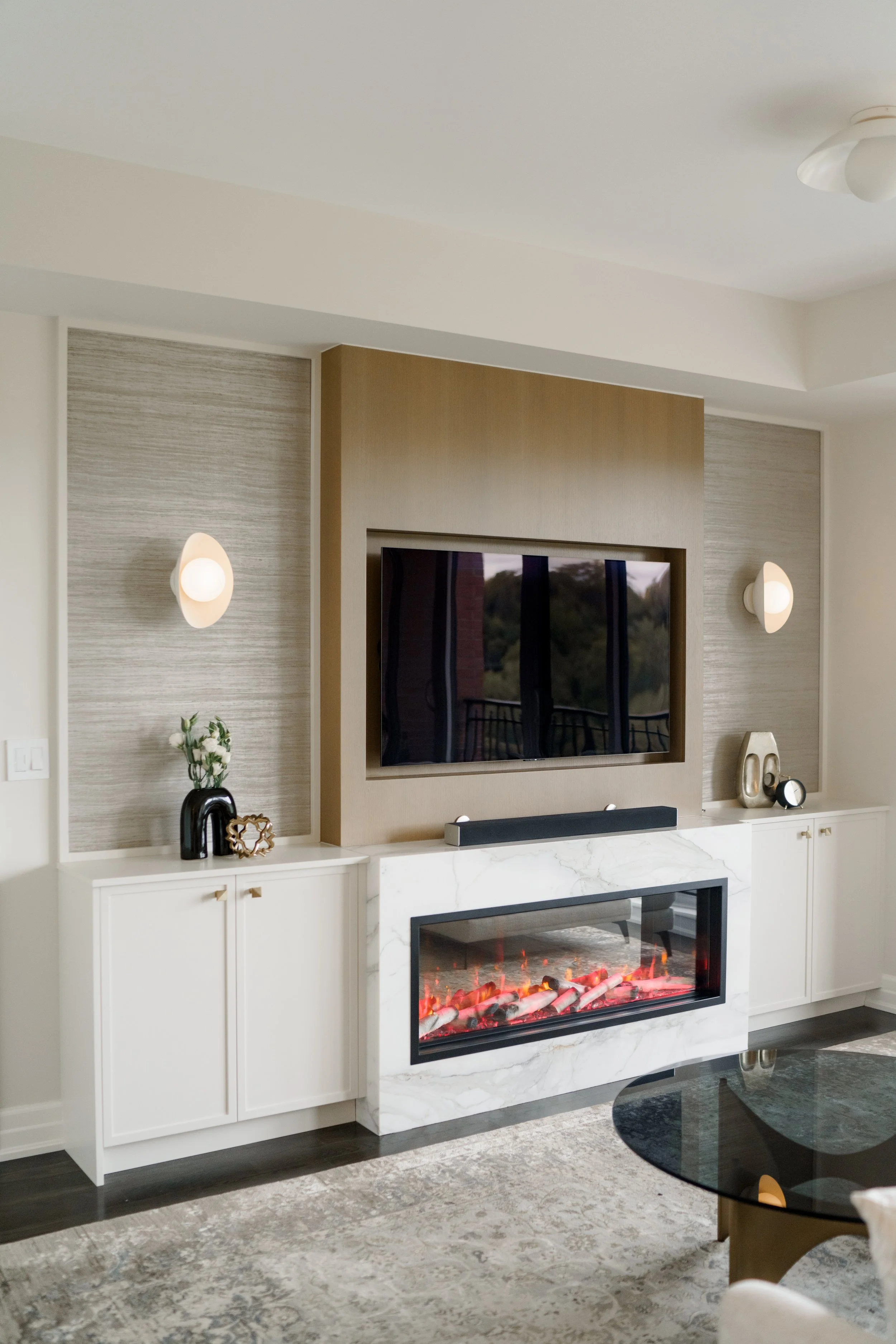 Living room with mounted TV, decorative pillows, white cabinetry with vases, a modern electric fireplace, beige and gray wall paneling, wall sconces, and a glass top coffee table.