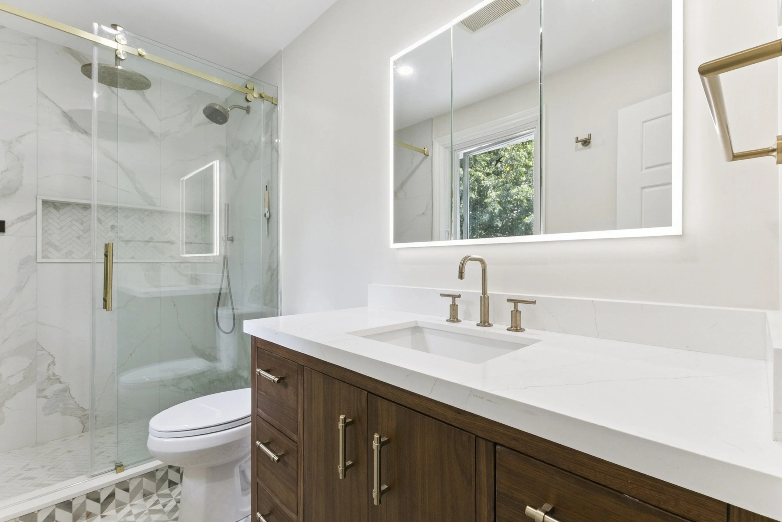 Modern bathroom with a glass-enclosed shower, white marble walls, wooden vanity with a white marble countertop, and a large backlit mirror. Window with trees visible outside.