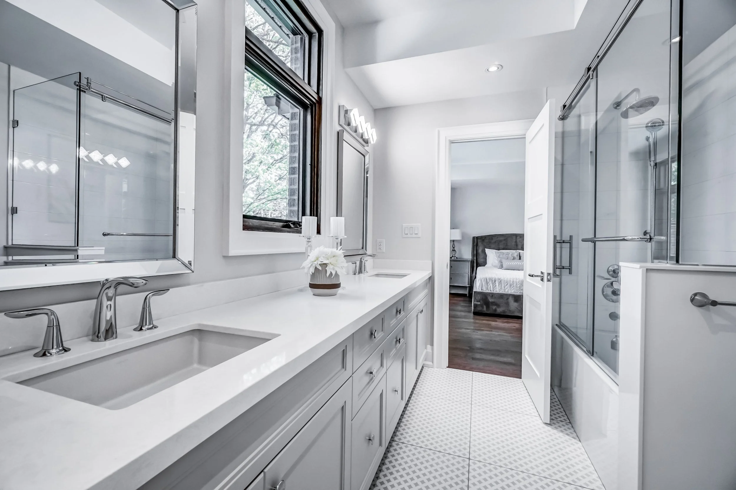 Modern bathroom with white vanity, double sink, gray cabinets, framed mirror, window, shower with glass doors, and a bedroom visible through open door.