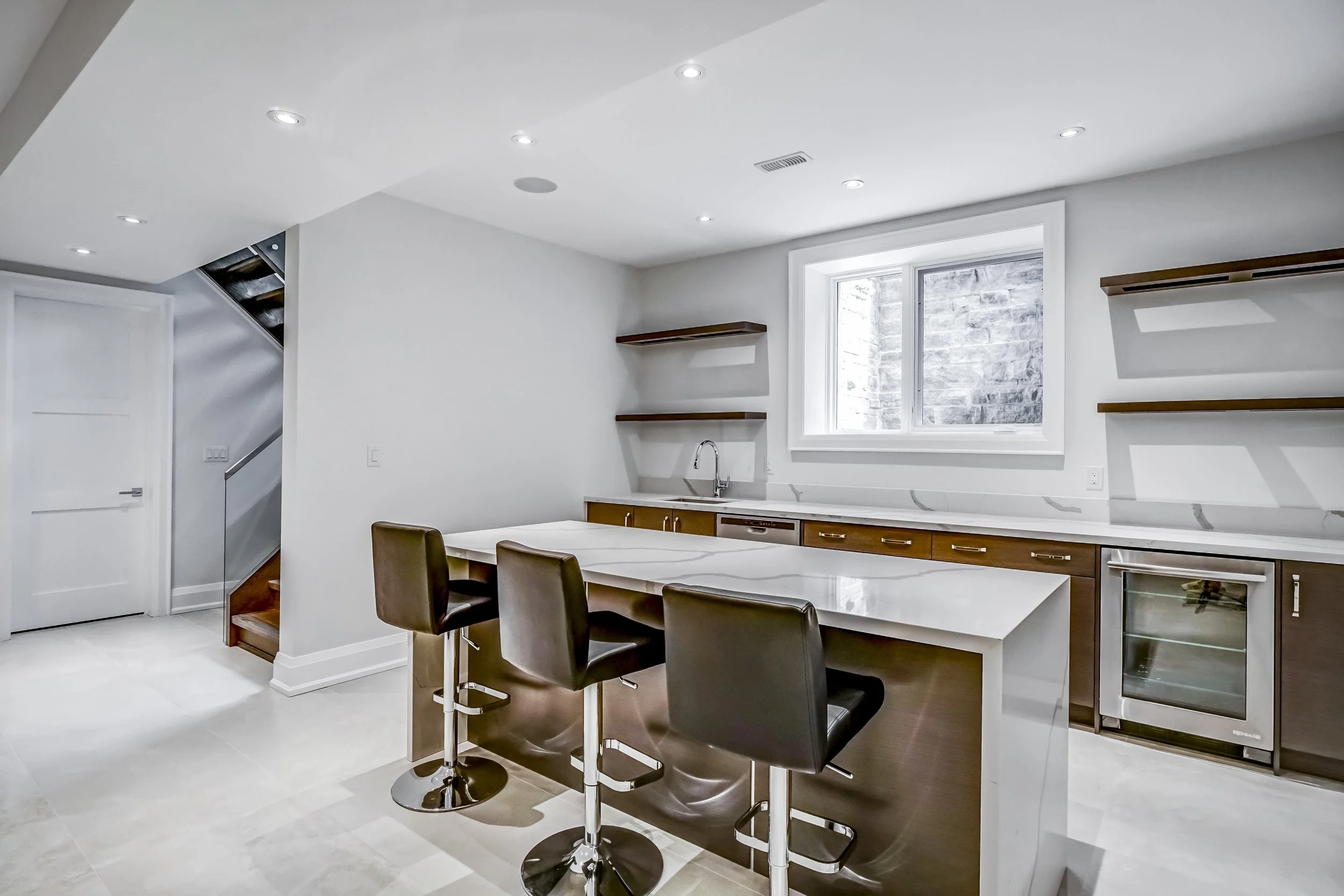 Modern kitchen with white walls, a large window, wooden shelves, a marble countertop, black barstools, and built-in appliances.
