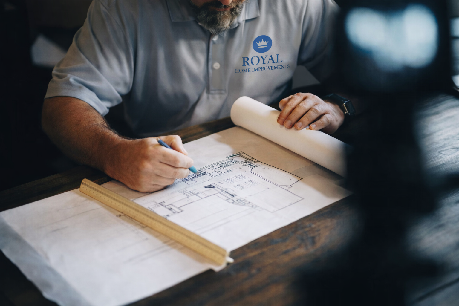 A man in a gray shirt with a 'Royal Home Improvements' logo is drawing on architectural blueprints with a blue pen, with a wooden ruler on the table.