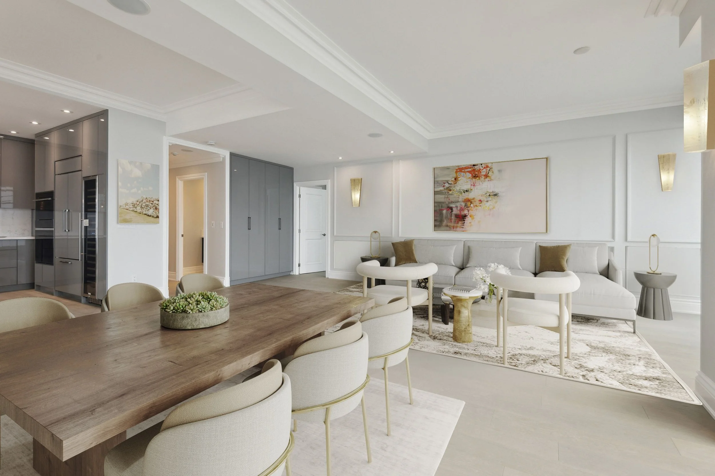 Open-concept living room and dining area in a modern home with white walls, light hardwood floors, a large wooden dining table with six chairs, a white sectional sofa with cushions, a colorful abstract painting on the wall, elegant wall sconces, and a kitchen area with stainless steel appliances and grey cabinetry.