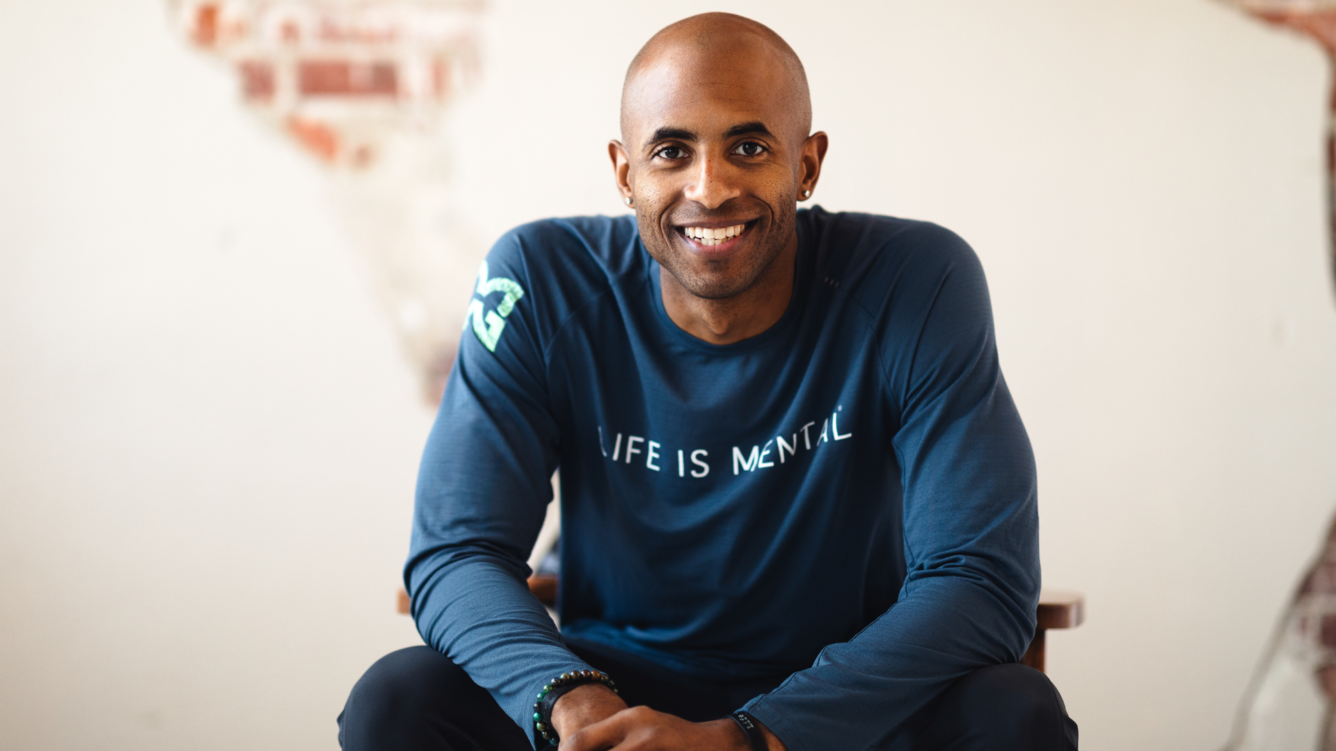 A smiling man with a shaved head sitting on a chair, wearing a navy blue long-sleeve shirt with the words "LIFE IS MENTAL" printed on it, indoors with a partially mural painted brick wall in the background.