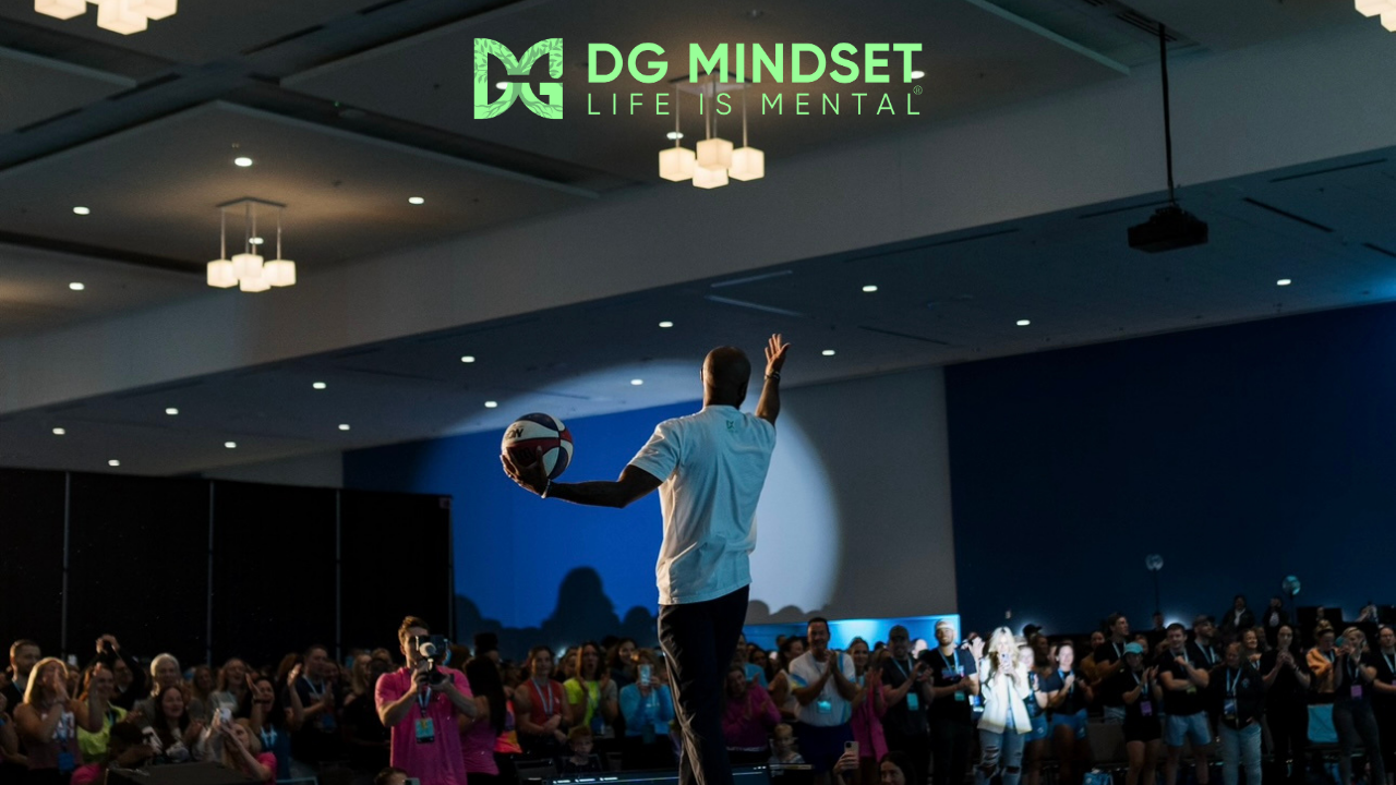 A speaker on stage holding a volleyball in one hand, addressing a large audience in a conference hall. The audience is clapping and taking photos, with a logo saying 'DG MINDSET LIFE IS MENTAL' at the top of the image.
