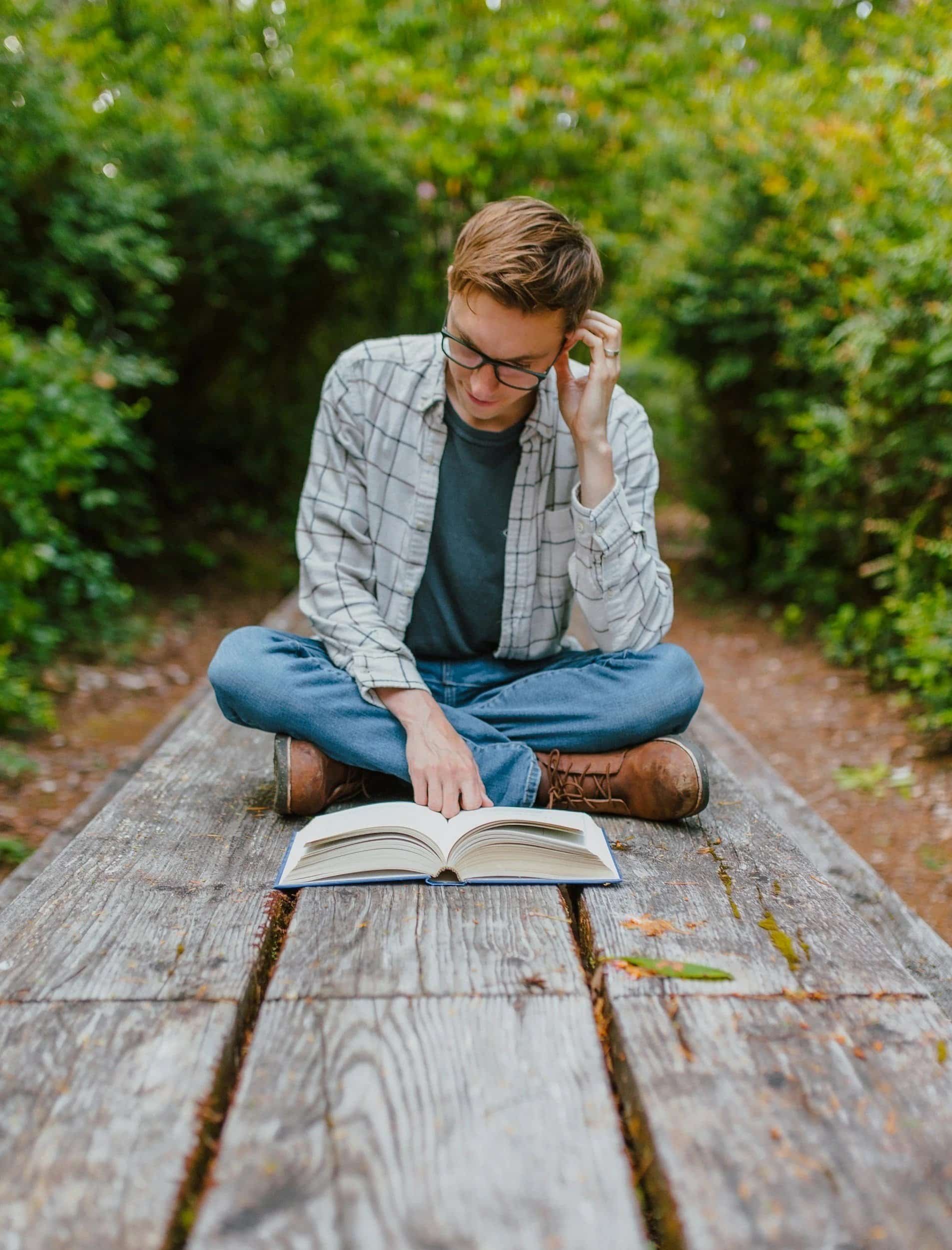A person with short brown hair and glasses sitting cross-legged on a wooden bench outdoors, reading a large open book. The person is wearing a plaid shirt over a dark t-shirt, and the background is filled with green foliage.