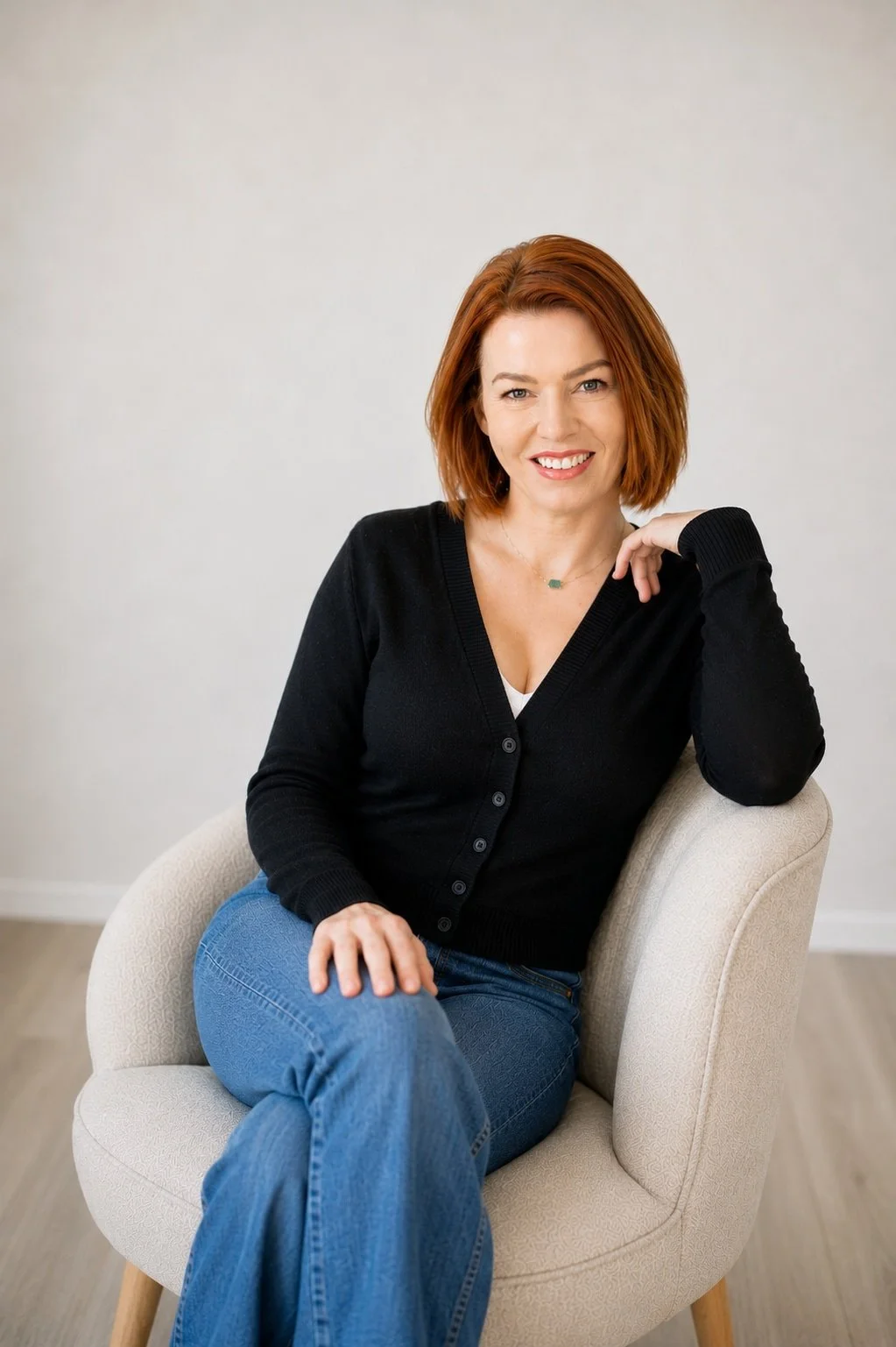 Tana with red hair in a black cardigan and blue jeans sits on a light chair, smiling warmly—an IFS EMDR therapist.