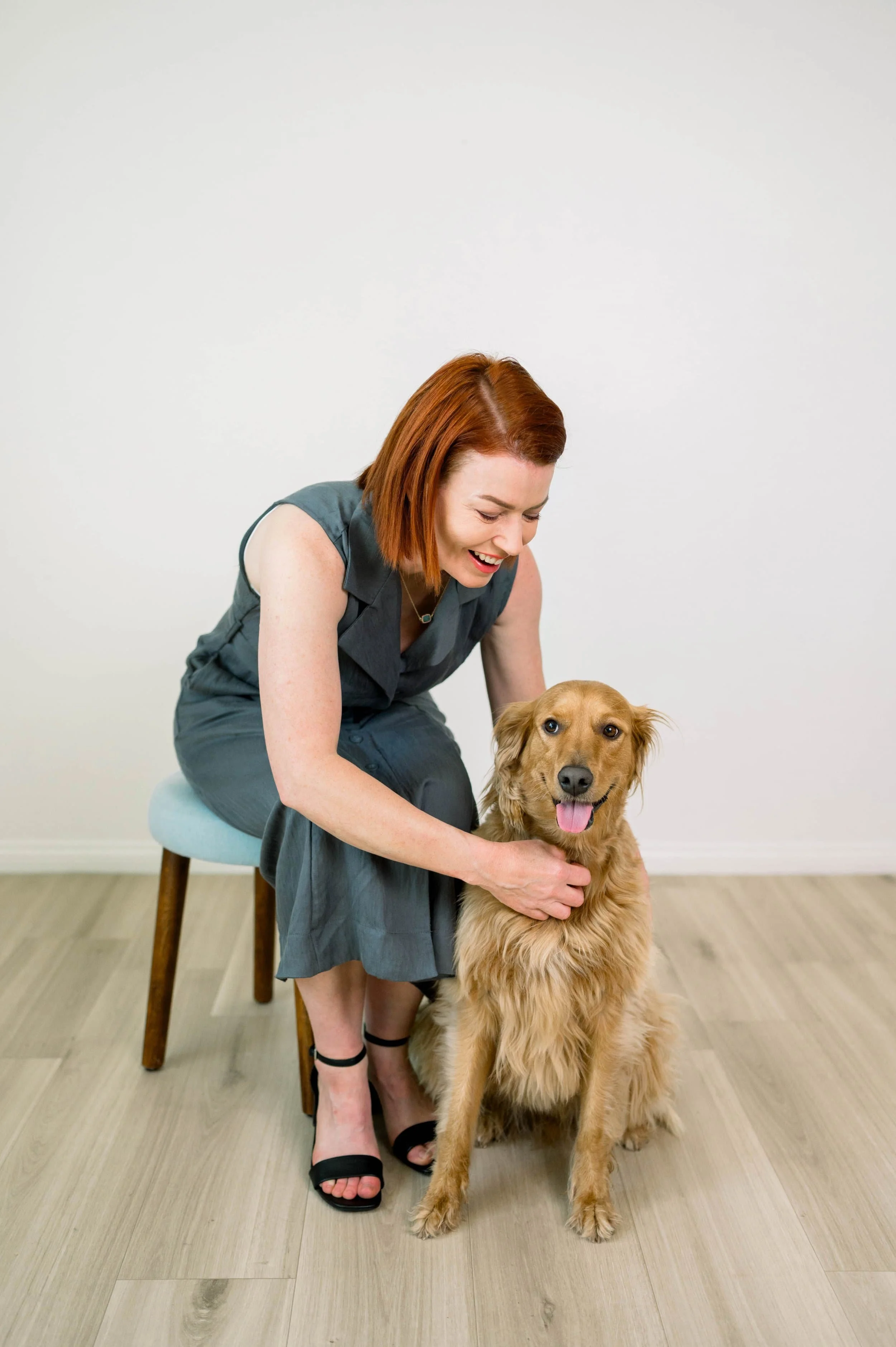 A woman with red hair smiling and petting a golden retriever dog sitting on the floor inside a room with light-colored wood flooring and a plain white wall.