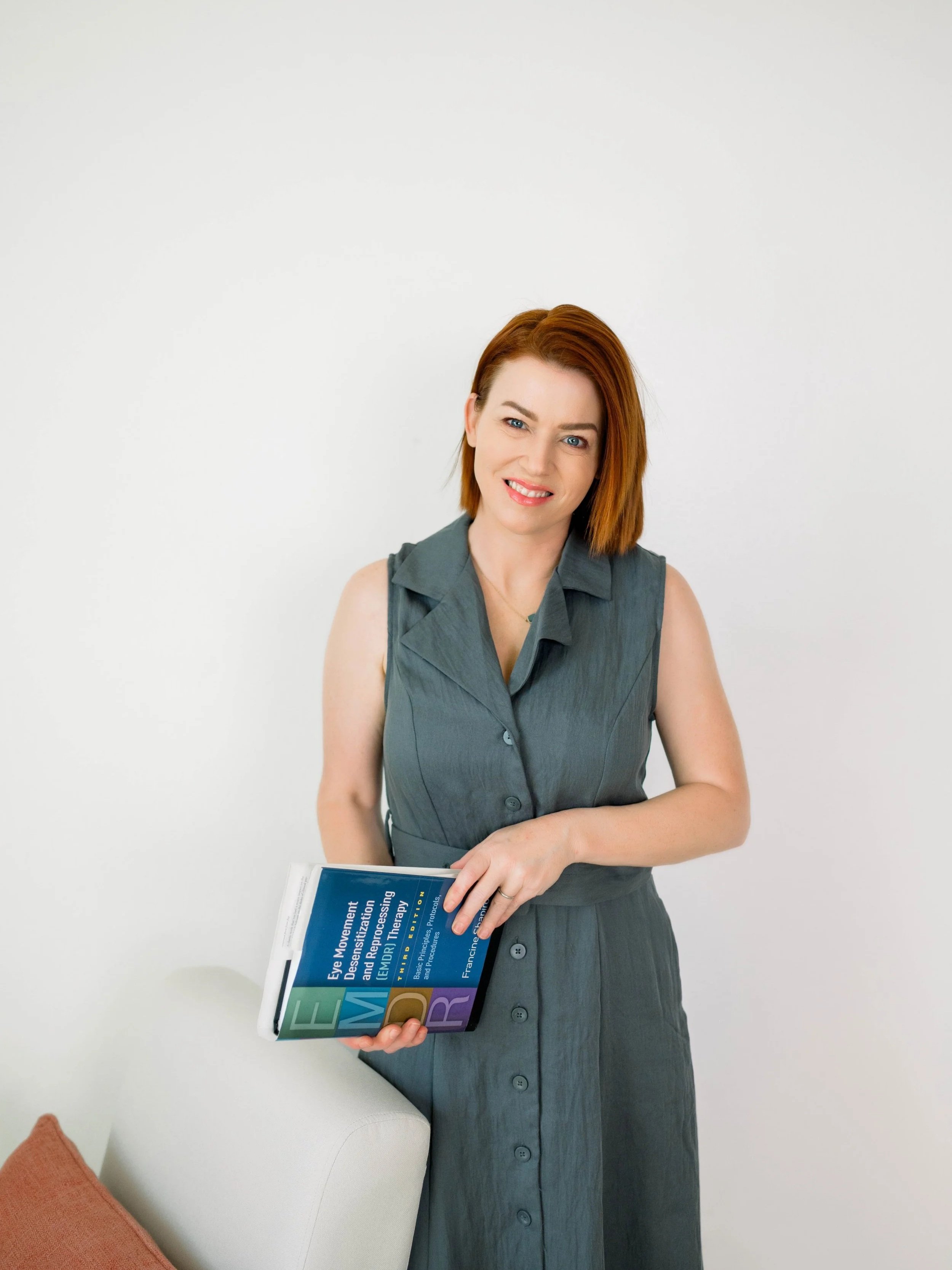 Tana Noonan with red hair and light skin is smiling at the camera while holding a book. She is wearing a sleeveless gray dress and standing against a plain white background.