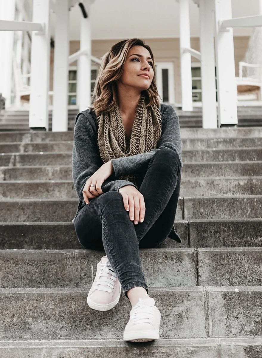A woman with shoulder-length hair sitting on outdoor concrete stairs, wearing a dark gray sweater, black jeans, white sneakers, and a large knitted scarf, looking off to the side.