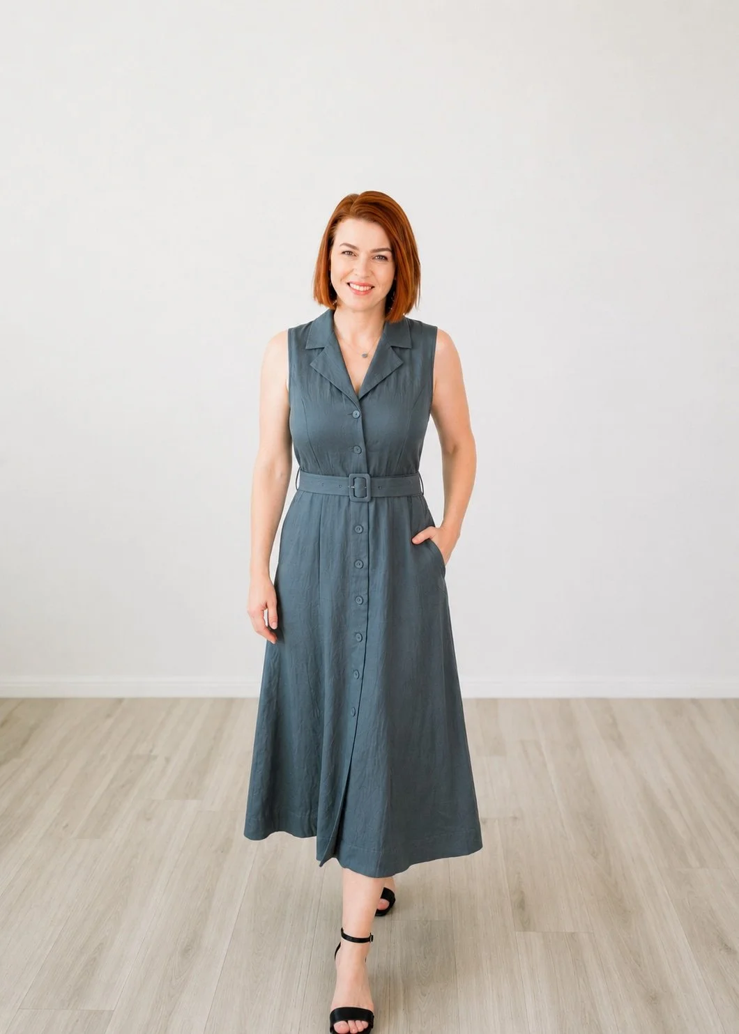 Tana with short red hair in a blue-gray dress and black sandals stands indoors, exuding calm perfect for a therapy setting.