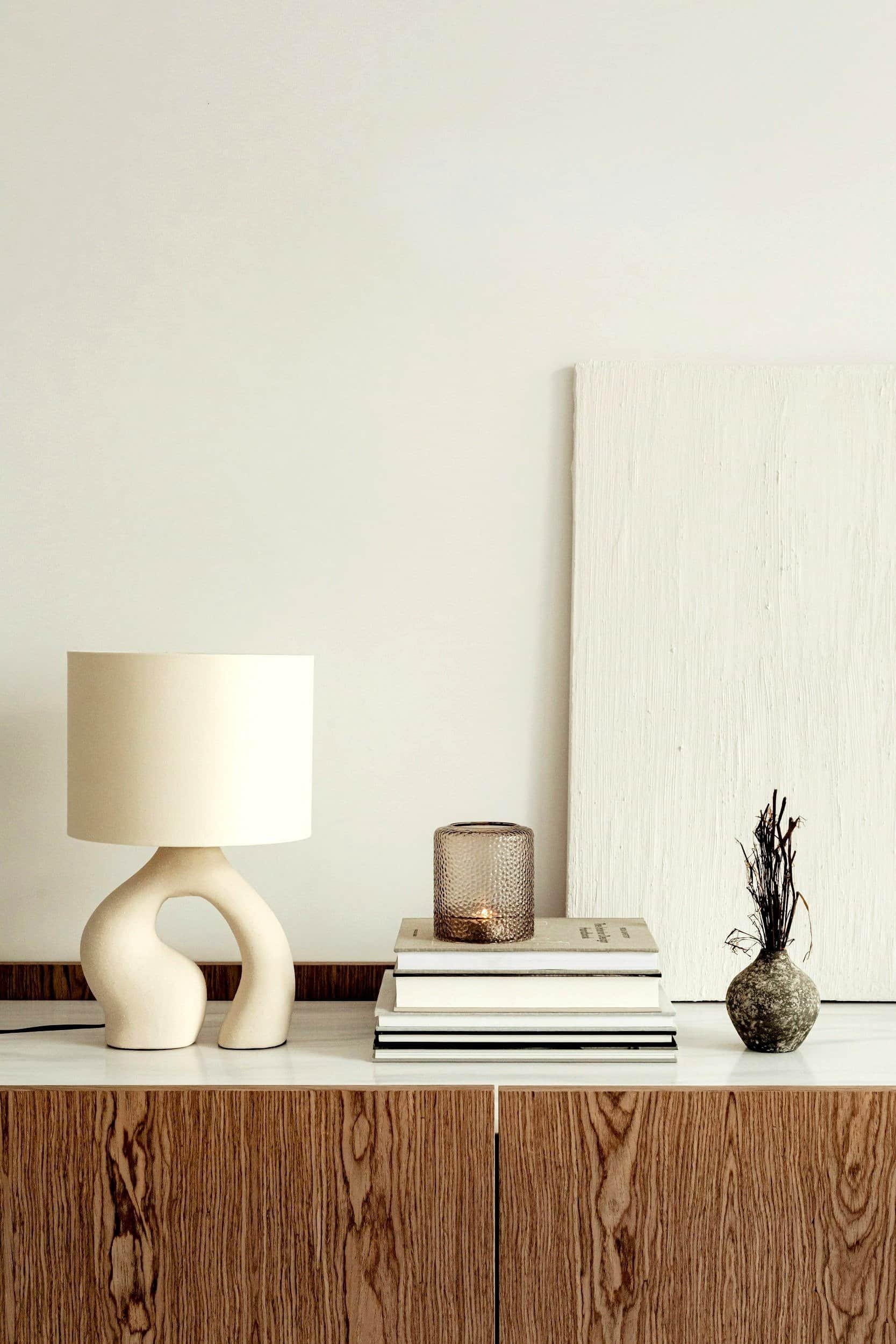A wooden sideboard with a white table lamp, a stack of books, a candle holder, and a decorative branch in a vase.