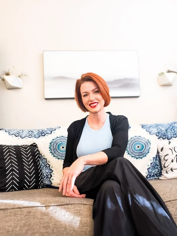 A woman with short red hair and red lipstick sitting on a sofa, smiling, with decorative pillows and wall art behind her.