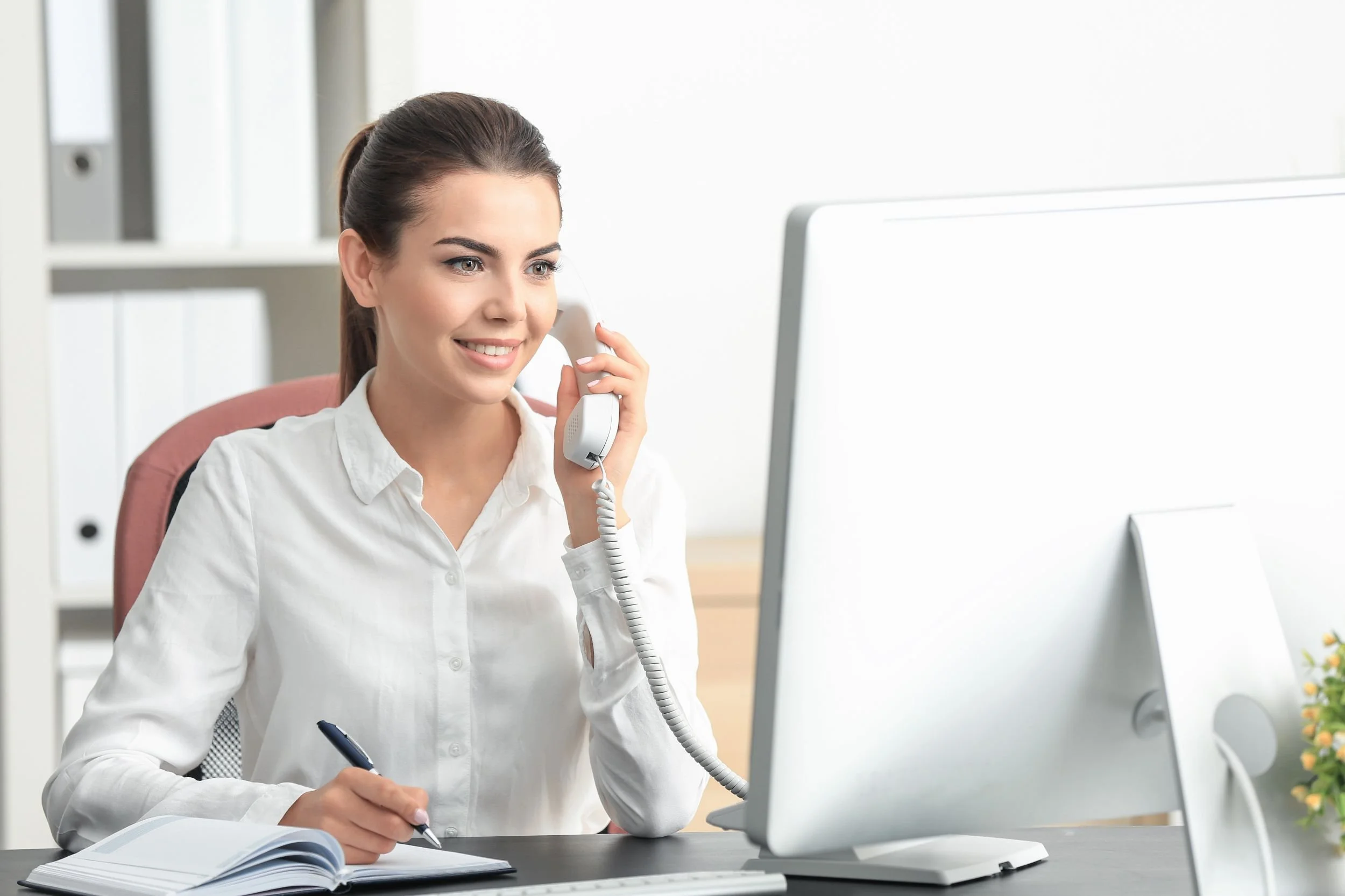 Young woman in white shirt smiling at computer while talking on phone with notebook on desk