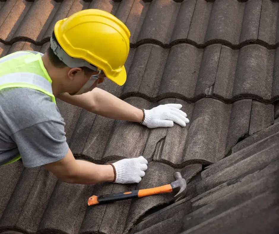 Roof maintenance worker in hard hat and safety vest replacing a damaged concrete tile during a routine roof inspection