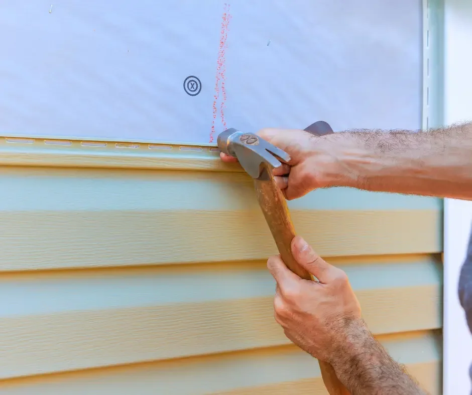 Close-up of GRM Roofing technician hammering a replacement beige vinyl siding panel into place in Ocean County, NJ