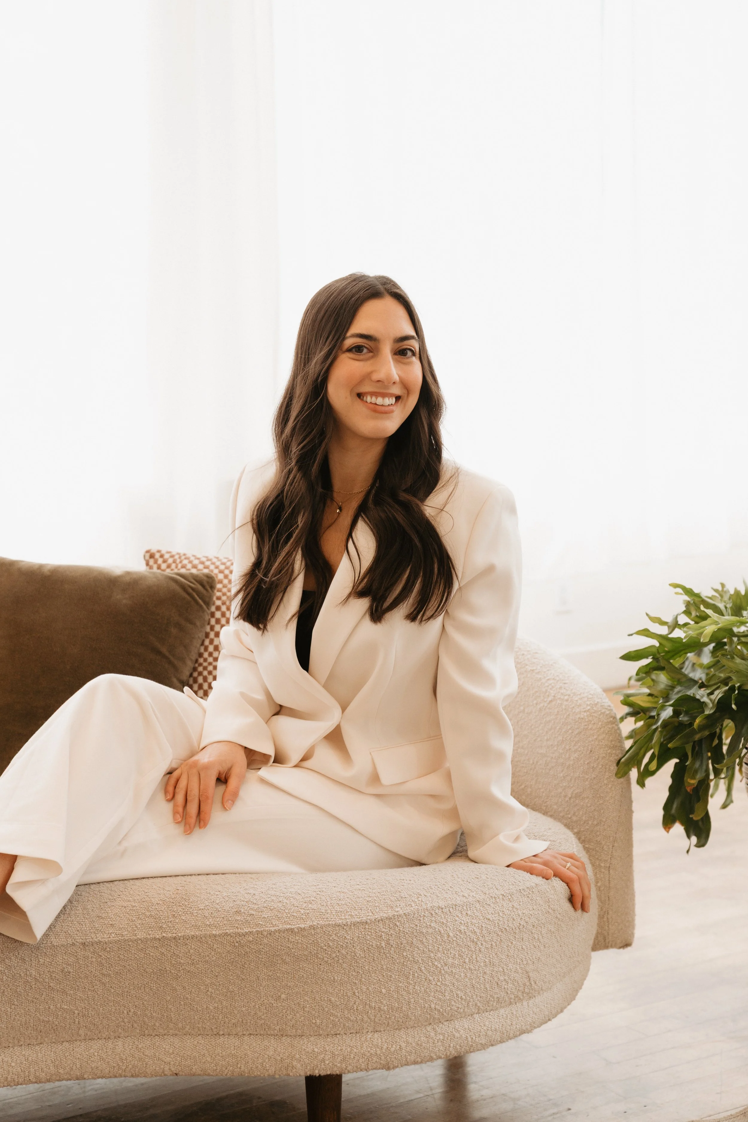 A woman with long dark hair, smiling, wearing a light-colored blazer and pants, sitting on a beige couch in a bright room with white curtains, a pillow, and a potted plant.