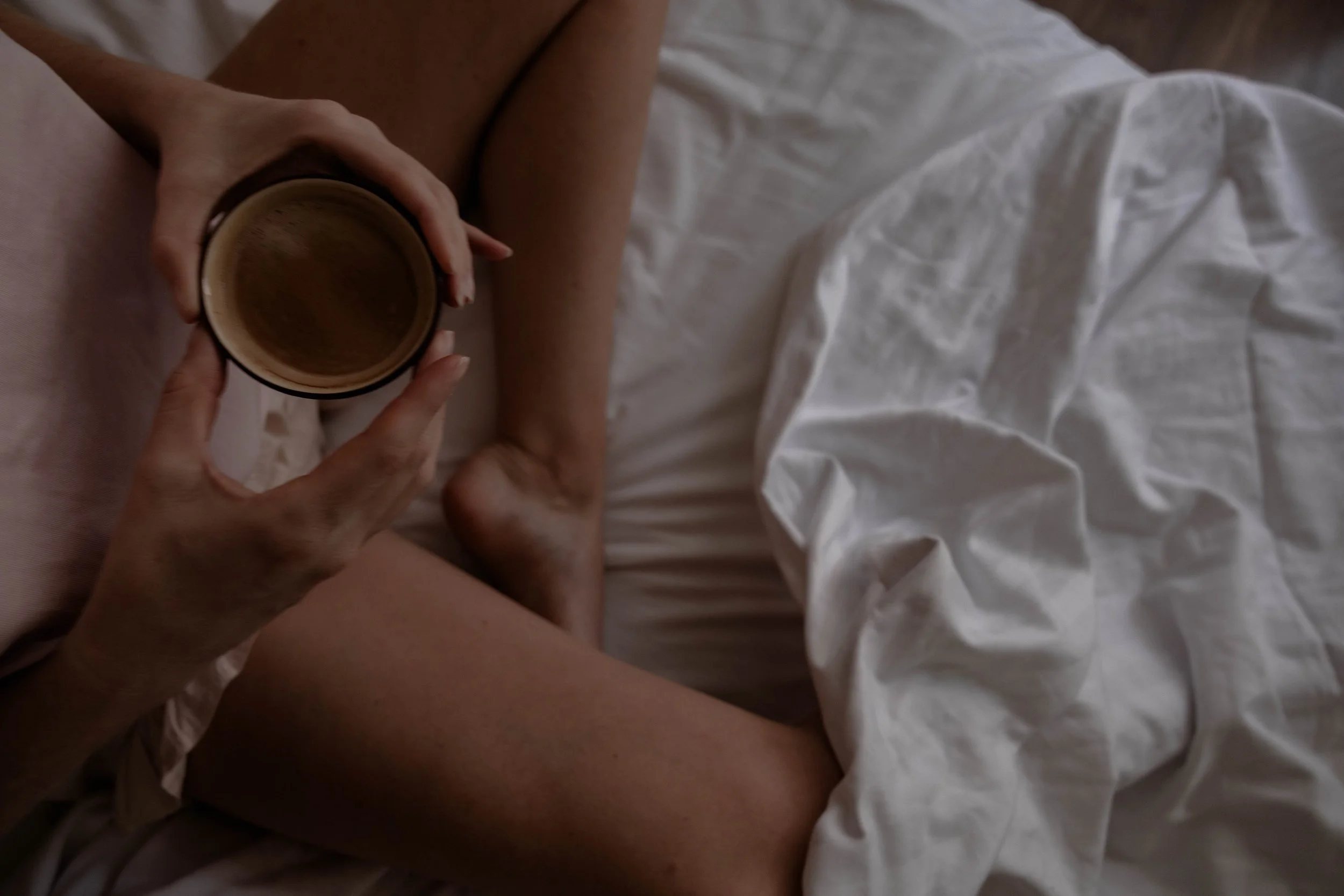 A person holding a cup of coffee while sitting on bed in a white shirt, with legs and white bedding visible.