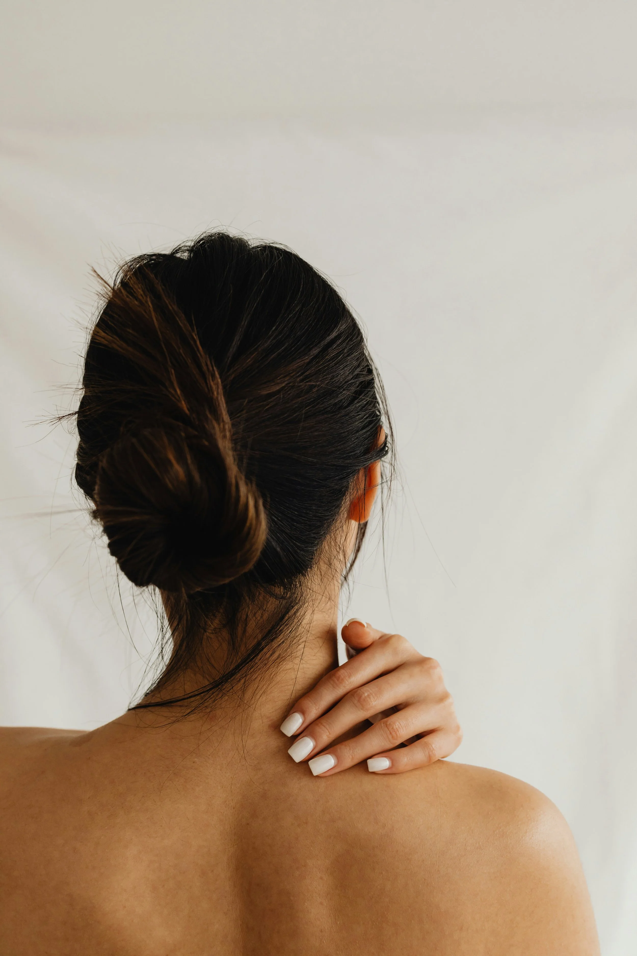 Back of a woman with dark brown hair styled in a bun, showing her bare shoulder and hand with white nail polish against a plain light background.