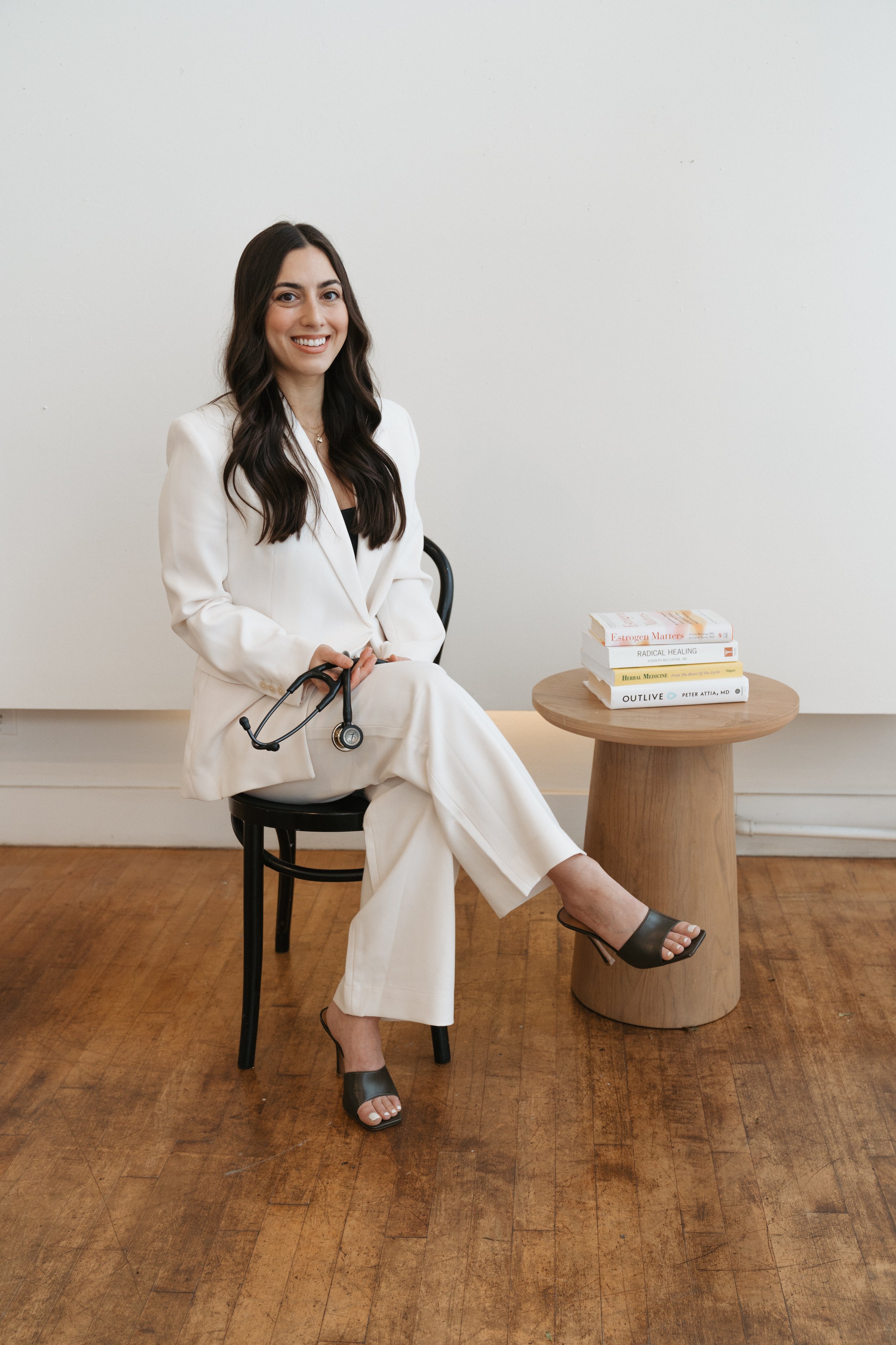 A woman with long dark hair, wearing a white suit and black high-heeled sandals, sitting on a black chair, holding a stethoscope, in a room with a white wall and wooden floor. There is a small round wooden table beside her with books on health and medicine.