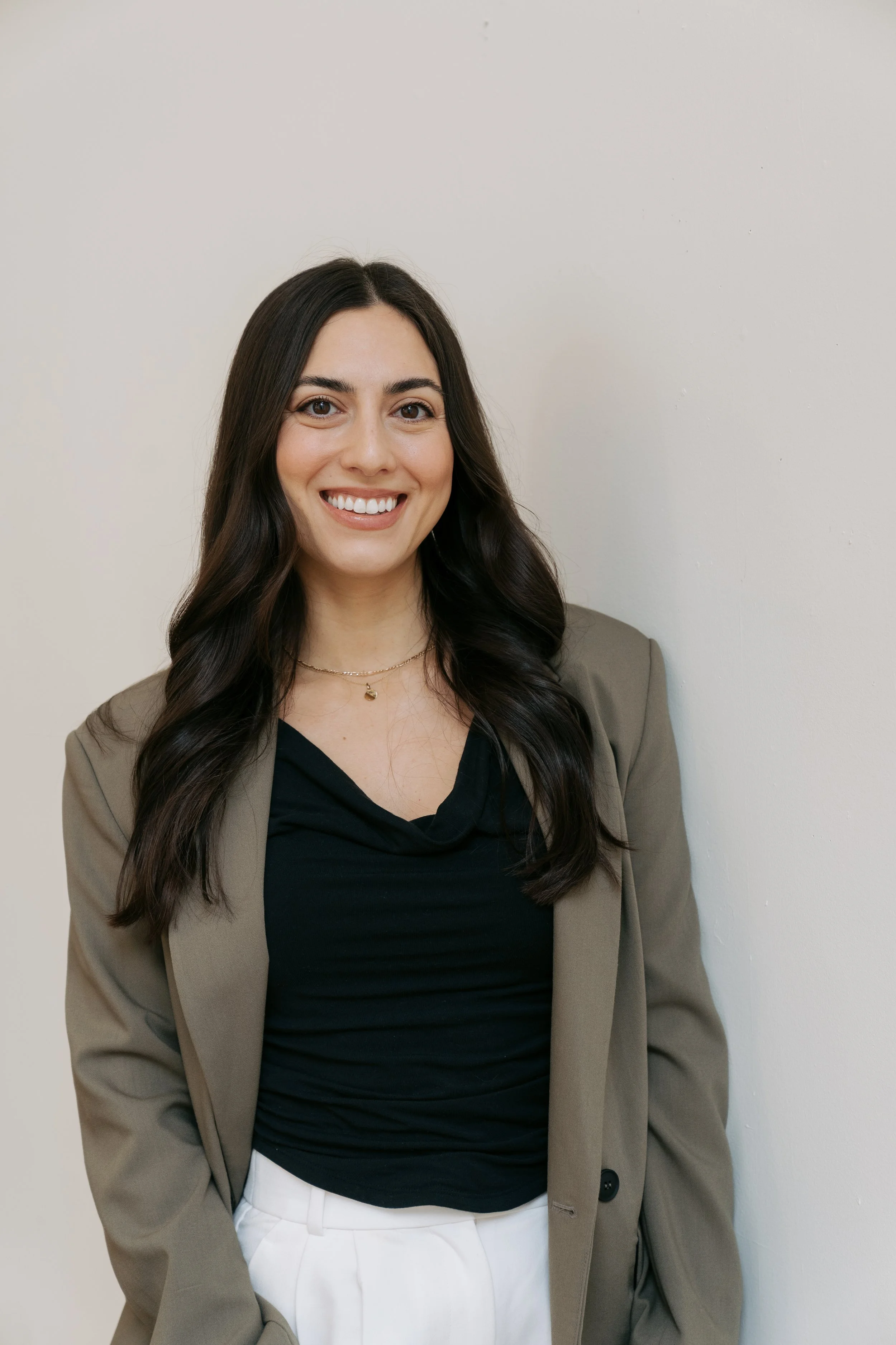 A woman with dark brown hair, smiling, wearing a beige blazer, black top, and white pants, standing against a plain off-white wall.