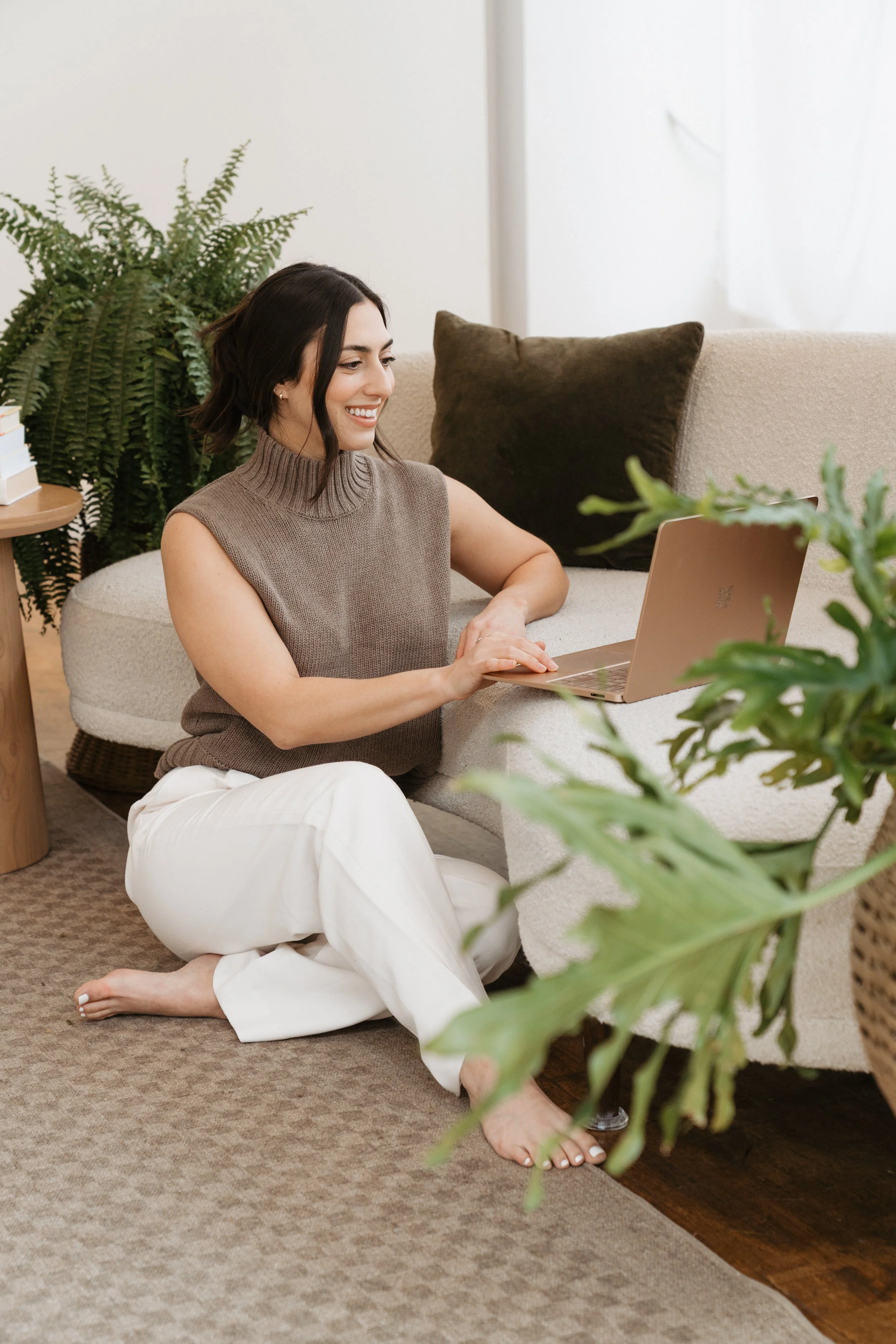 A woman sitting on the floor next to a beige sofa, working on a laptop. She has short dark hair and is wearing a sleeveless knit top and white pants. There are green plants and a brown pillow on the sofa.