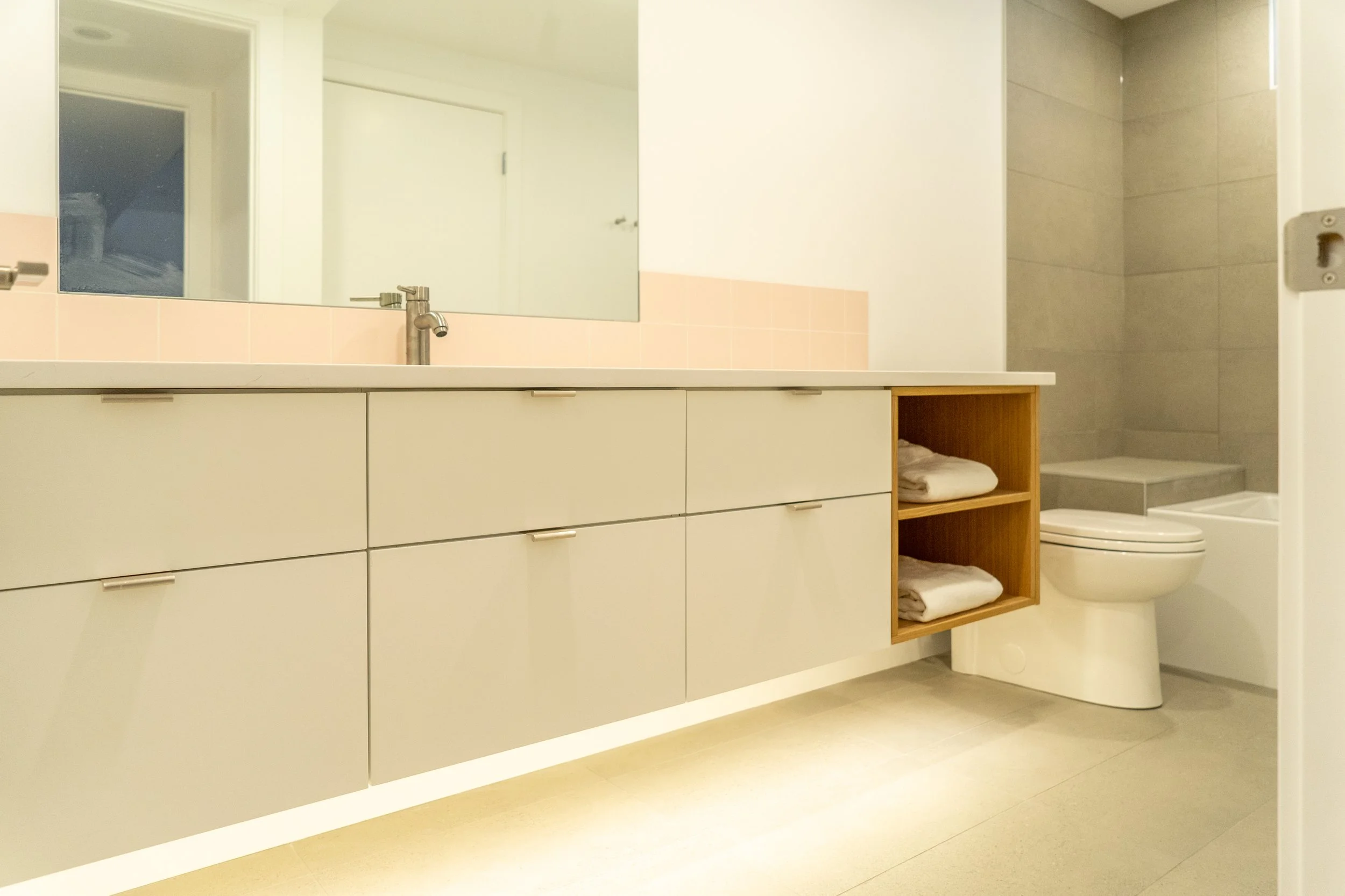 A modern bathroom featuring a long white vanity with multiple drawers and a stainless steel faucet, a large mirror, open wooden shelves with folded towels, a toilet, and a walk-in shower with beige tiles.