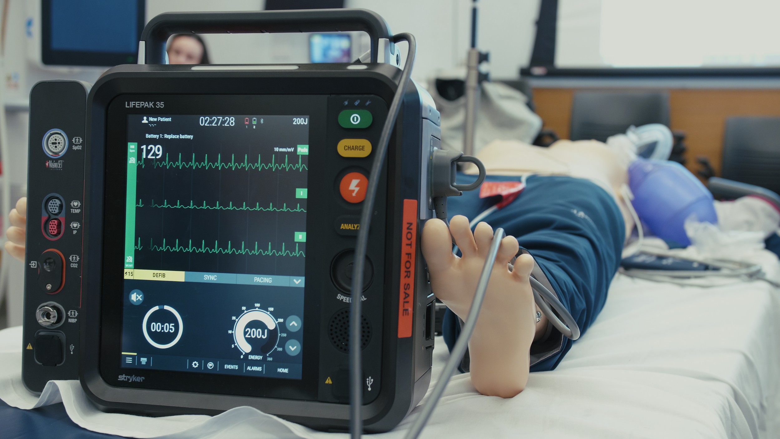 Illustration of a defibrillator on a medical cart in a hospital room, with cables and storage boxes.