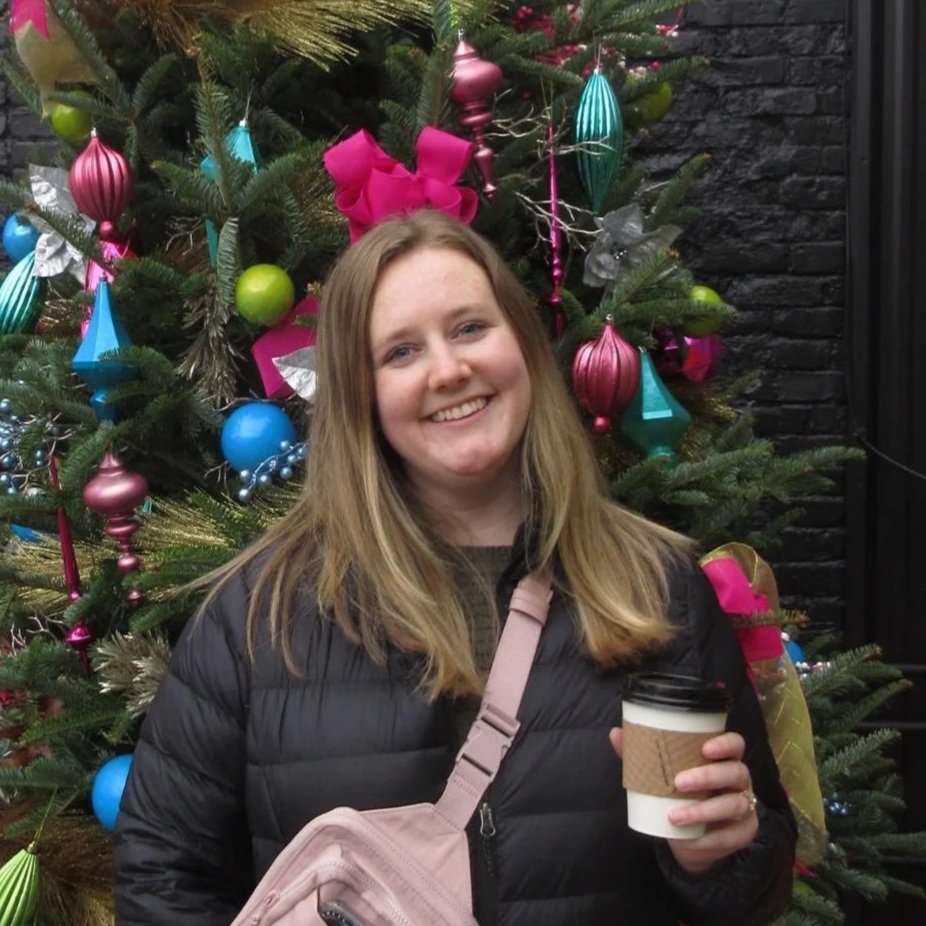 A young woman smiles at the camera. She is in front of a Christmas tree with colorful decorations. She has long, blond hair, is holding a hot coffee, has a pink shoulder bag, and is wearing a black puffer jacket.